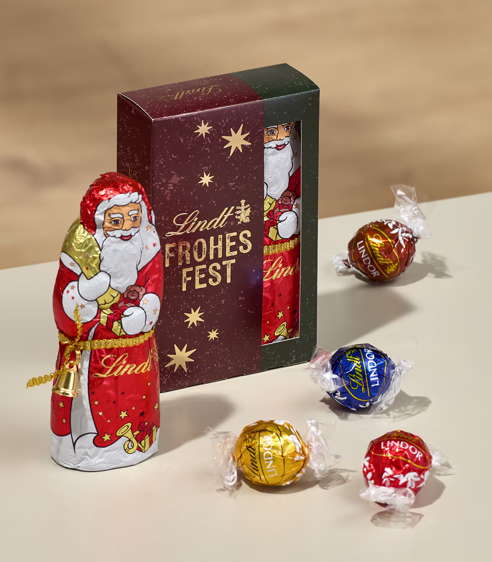 Im Vordergrund stehen eine Schokoladenfigur des Weihnachtsmanns und mehrere verpackte Lindor-Pralinen vor einer festlichen Lindt-Geschenkbox vor einem schlichten beigen Hintergrund. (Beschreibungstext KI-generiert)