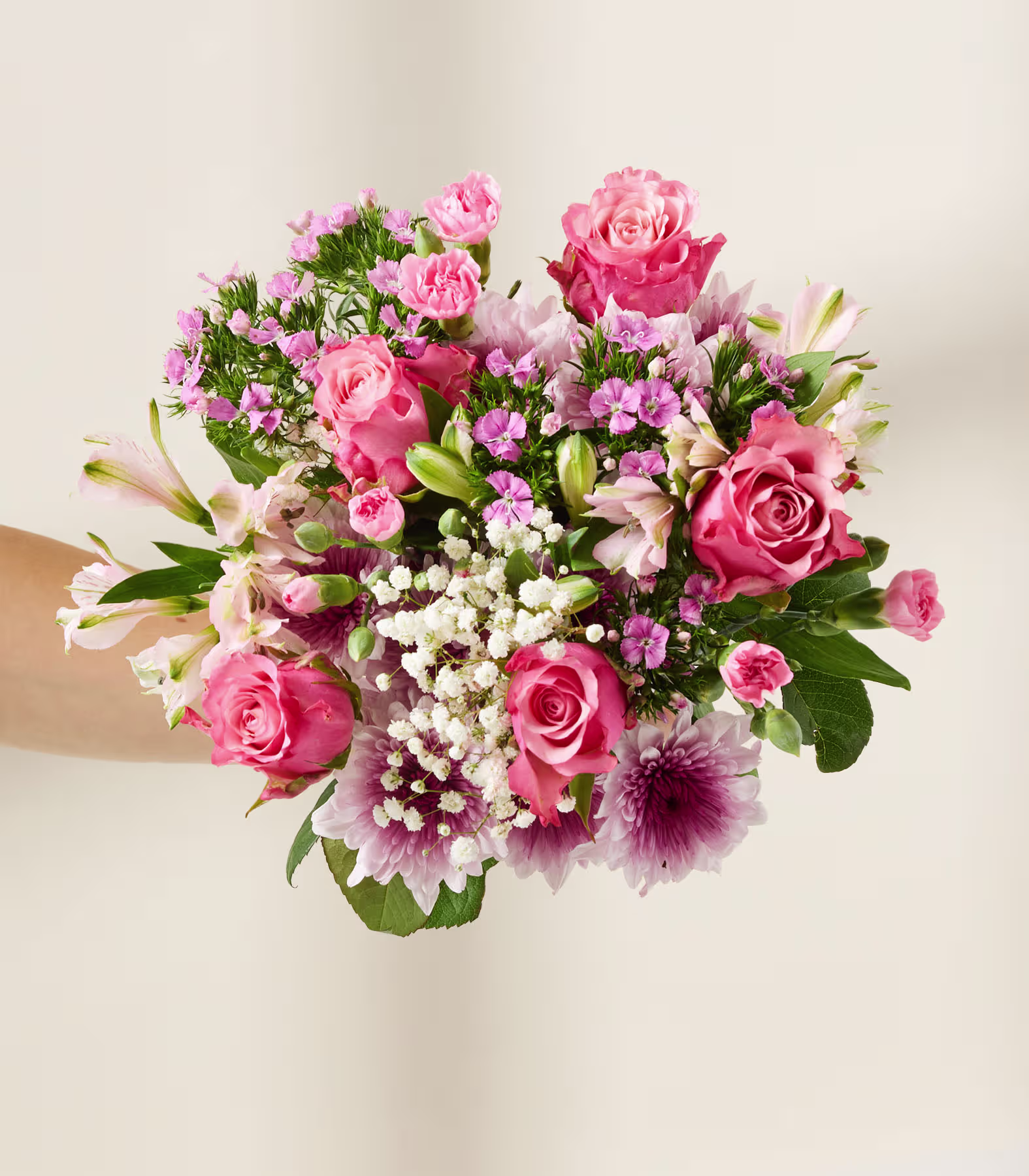 Ein lebhafter Strauß aus rosa Rosen, weißen Nelken und anderen farbenfrohen Blumen, arrangiert in einer Vase, mit einer Hand, die den Strauß hält, vor einem schlichten beigen Hintergrund. (Beschreibungstext KI-generiert)