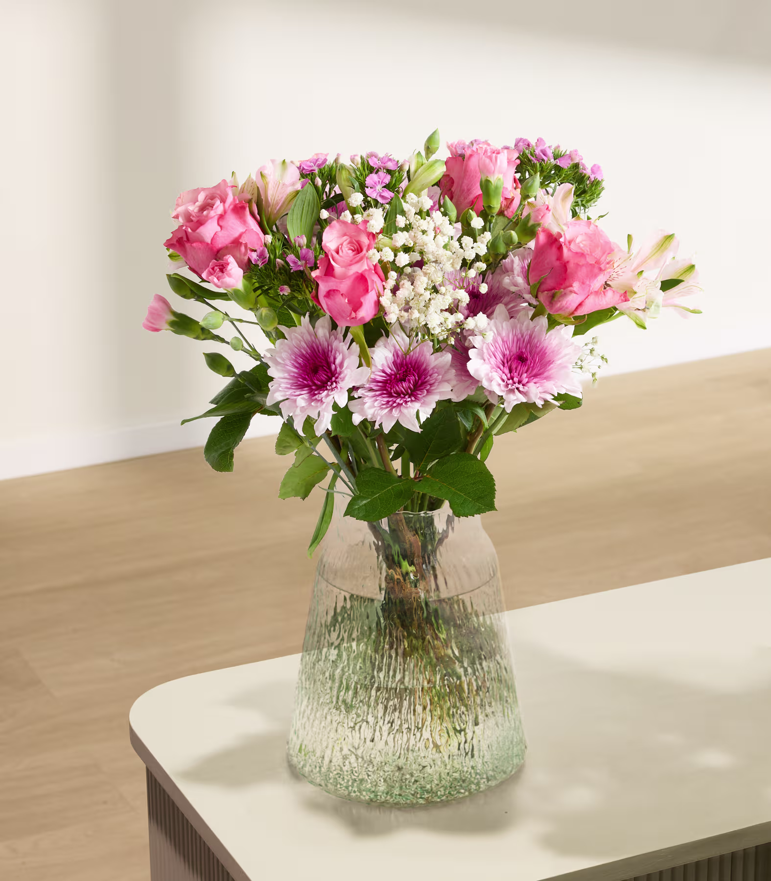 Ein wunderschöner Strauß leuchtender Blumen, darunter Nelken, Rosen und andere Blüten, steht in einer Glasvase auf einem Holztisch vor einem schlichten Hintergrund. (Beschreibungstext KI-generiert)