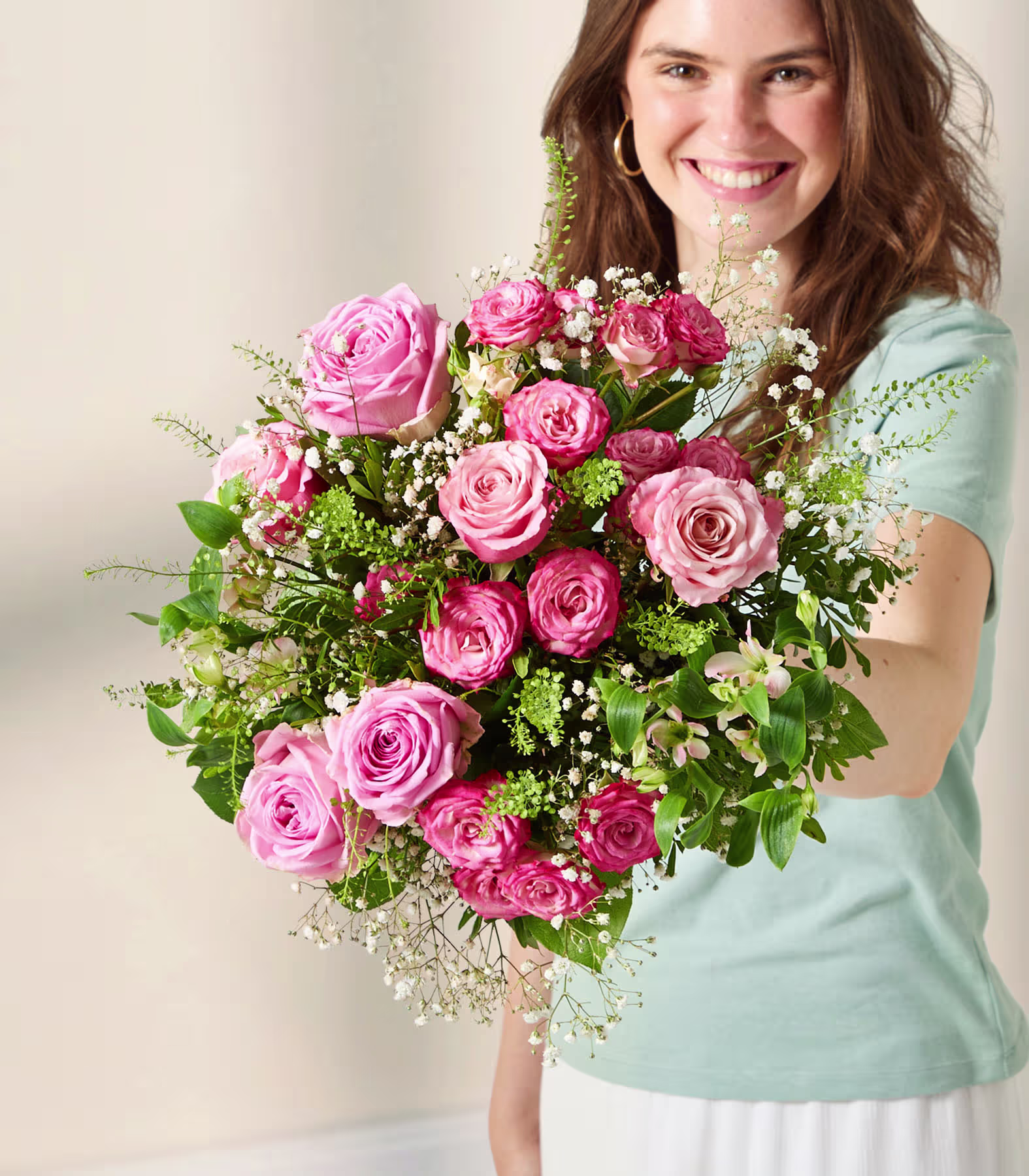 Auf einem Holztisch in einem gemütlichen Wohnzimmer steht ein Strauß leuchtend pinkfarbener Rosen und anderer Blumen; ein graues Sofa und ein großes Fenster sorgen für natürliches Licht. (Beschreibungstext KI-generiert)