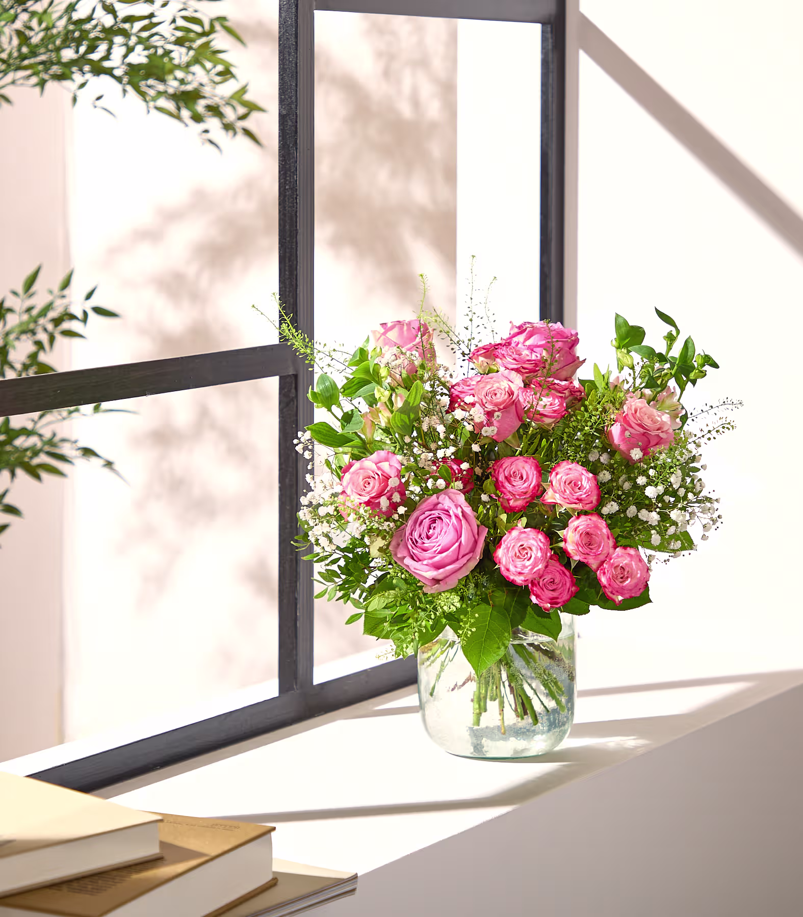 Ein Strauß rosa Rosen in einer Glasvase steht auf einem weißen Fensterbrett vor einem schwarzgerahmten Fenster. (Beschreibungstext KI-generiert)