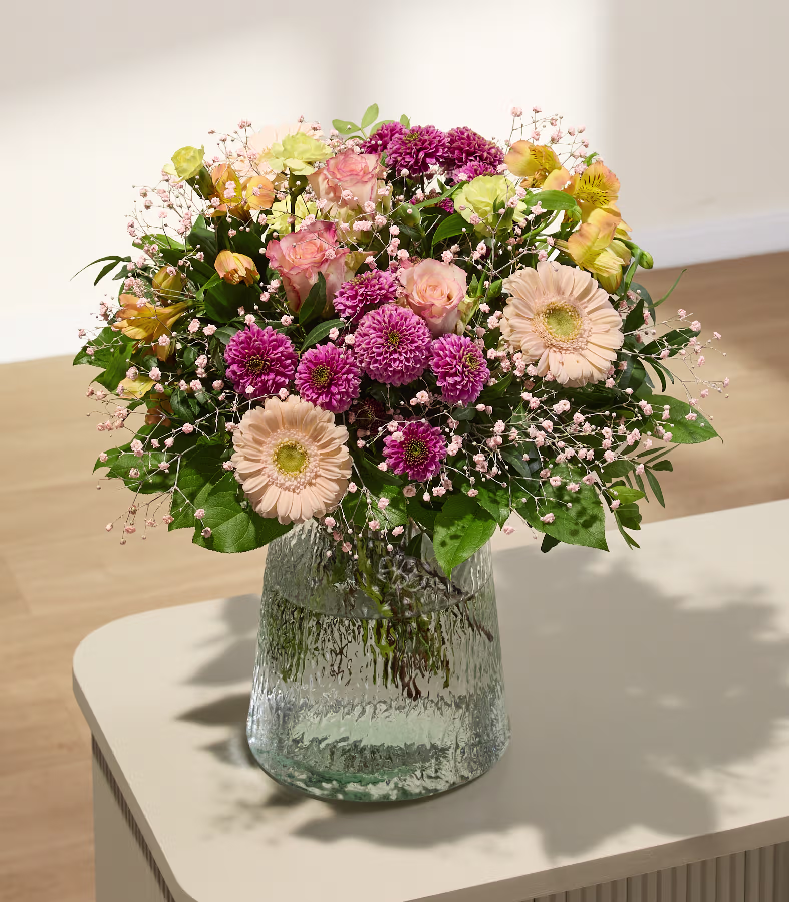 Ein farbenfroher Strauß aus rosa, pfirsichfarbenen und gelben Blumen füllt eine strukturierte Glasvase auf einem hellen Tisch; im Hintergrund ist eine sanft beleuchtete, strukturierte Wand zu sehen. (Beschreibungstext KI-generiert)
