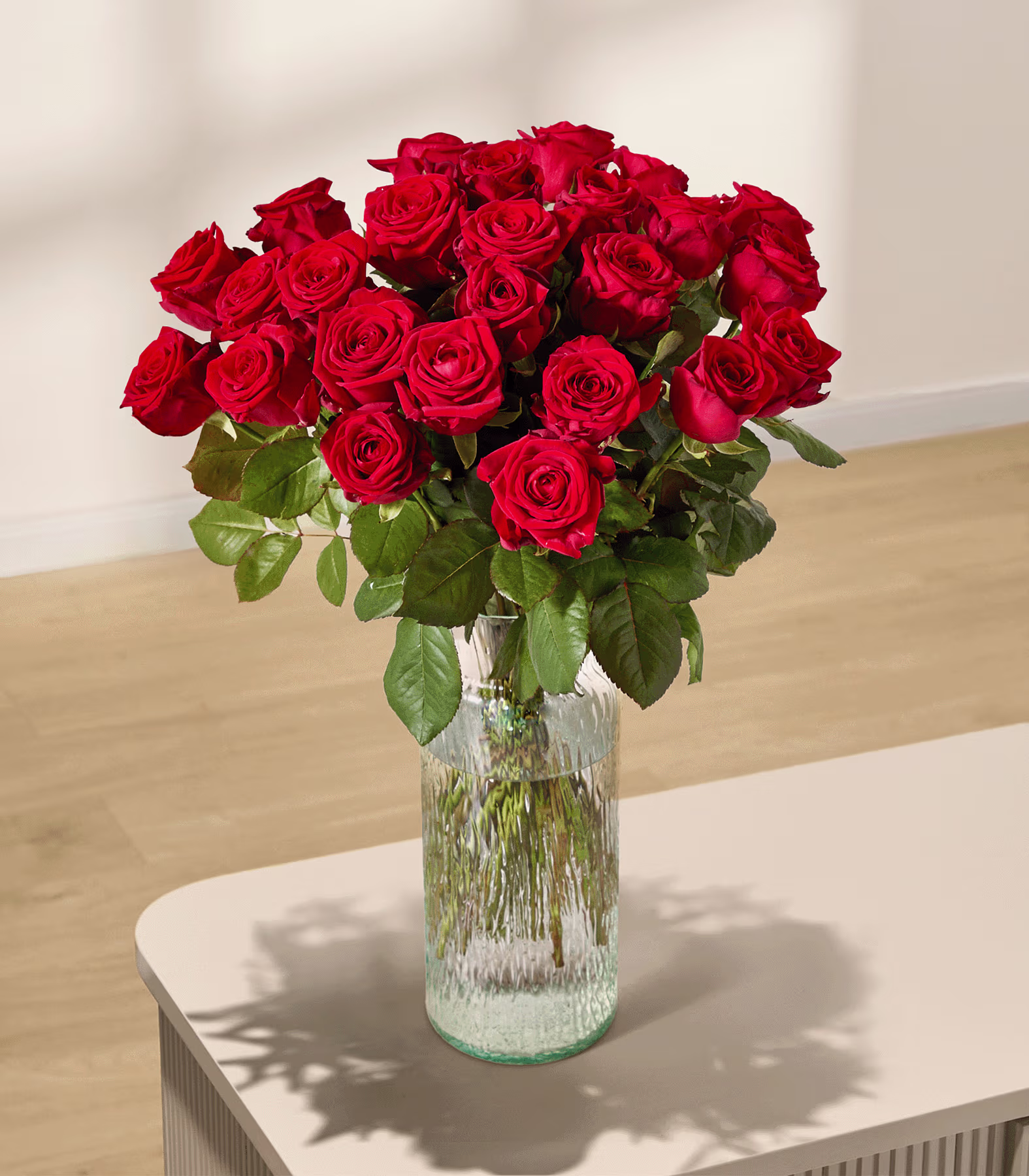 Auf einem hellen Tisch steht ein leuchtender Strauß roter Rosen in einer strukturierten Glasvase; im Hintergrund ist eine sanft beleuchtete Wand zu sehen. (Beschreibungstext KI-generiert)