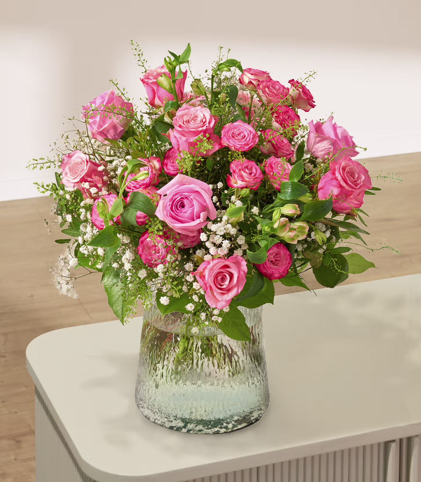 Ein üppiger Strauß aus rosa Rosen und Schleierkraut in einer strukturierten Glasvase steht auf einem weißen Tisch vor einer neutralen Wand. (Beschreibungstext KI-generiert)