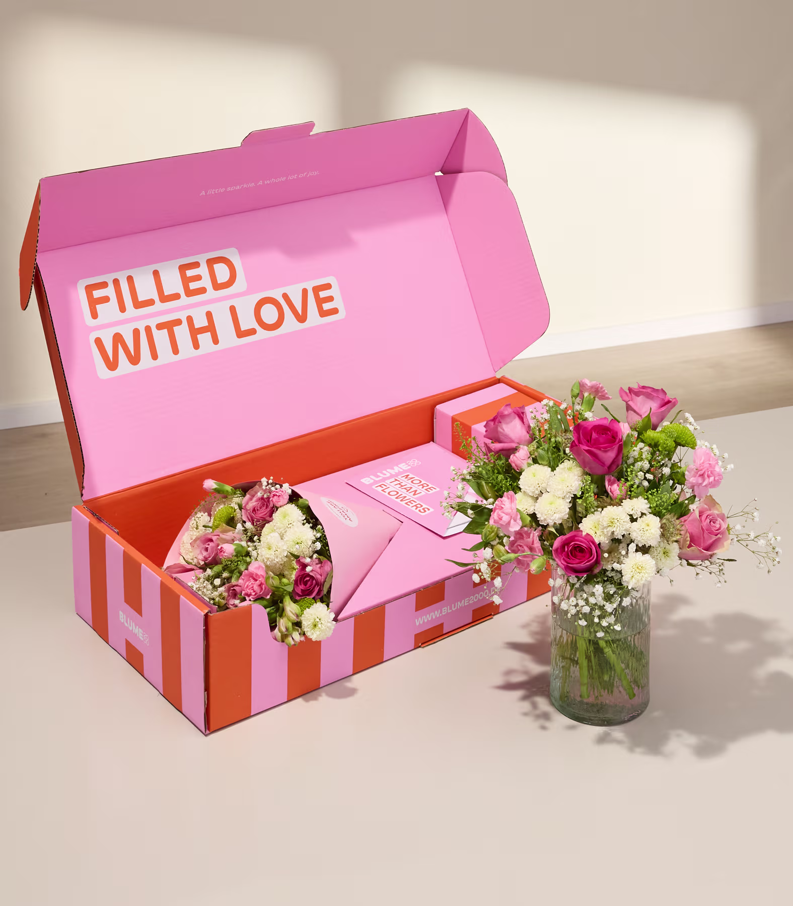 Eine rosa-orange gestreifte Geschenkbox mit Blumen und einer Karte steht neben einer Glasvase mit rosa und weißen Blüten vor einer schlichten, hellen Wand. (Beschreibungstext KI-generiert)