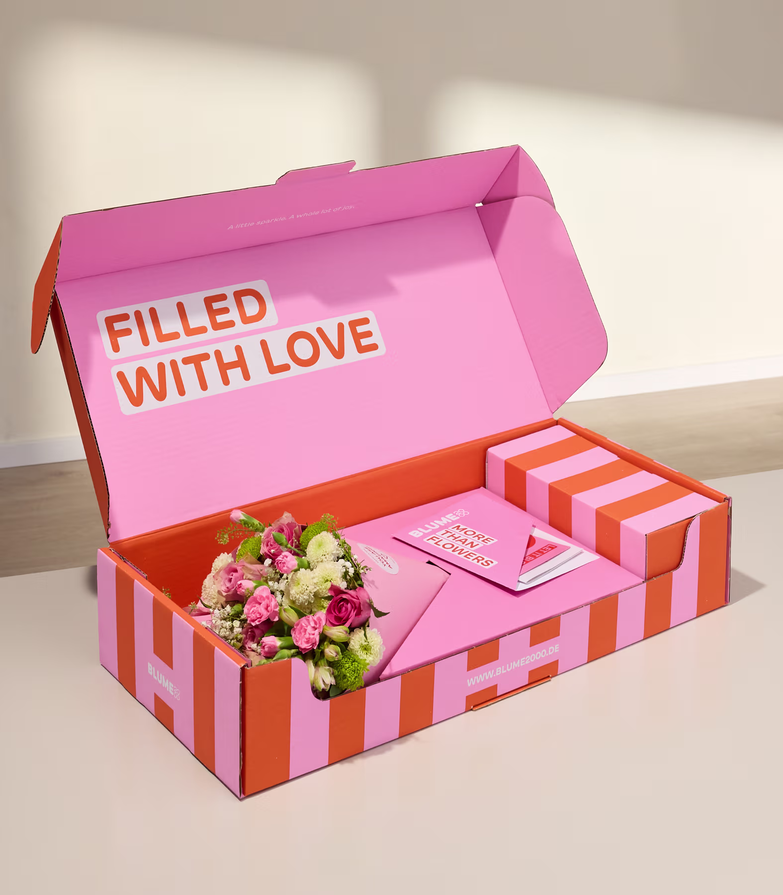 Eine rosa-orange gestreifte Geschenkbox mit einem Blumenstrauß und einer Karte steht auf einer schlichten, hellen Fläche vor einer neutralen Wand. (Beschreibungstext KI-generiert)