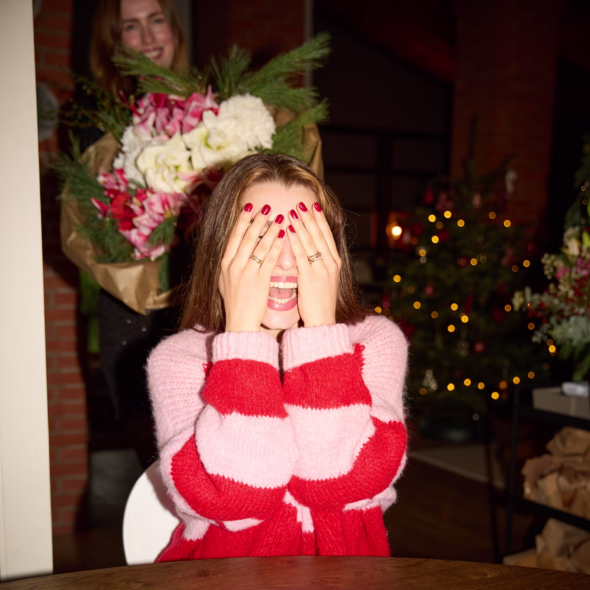 Im Vordergrund hält sich eine Frau in einem rot-rosa gestreiften Pullover die Augen zu, während sich von hinten eine andere Frau mit einem großen Blumenstrauß in einem schwach beleuchteten Raum mit einem Weihnachtsbaum nähert. (Beschreibungstext KI-generiert)