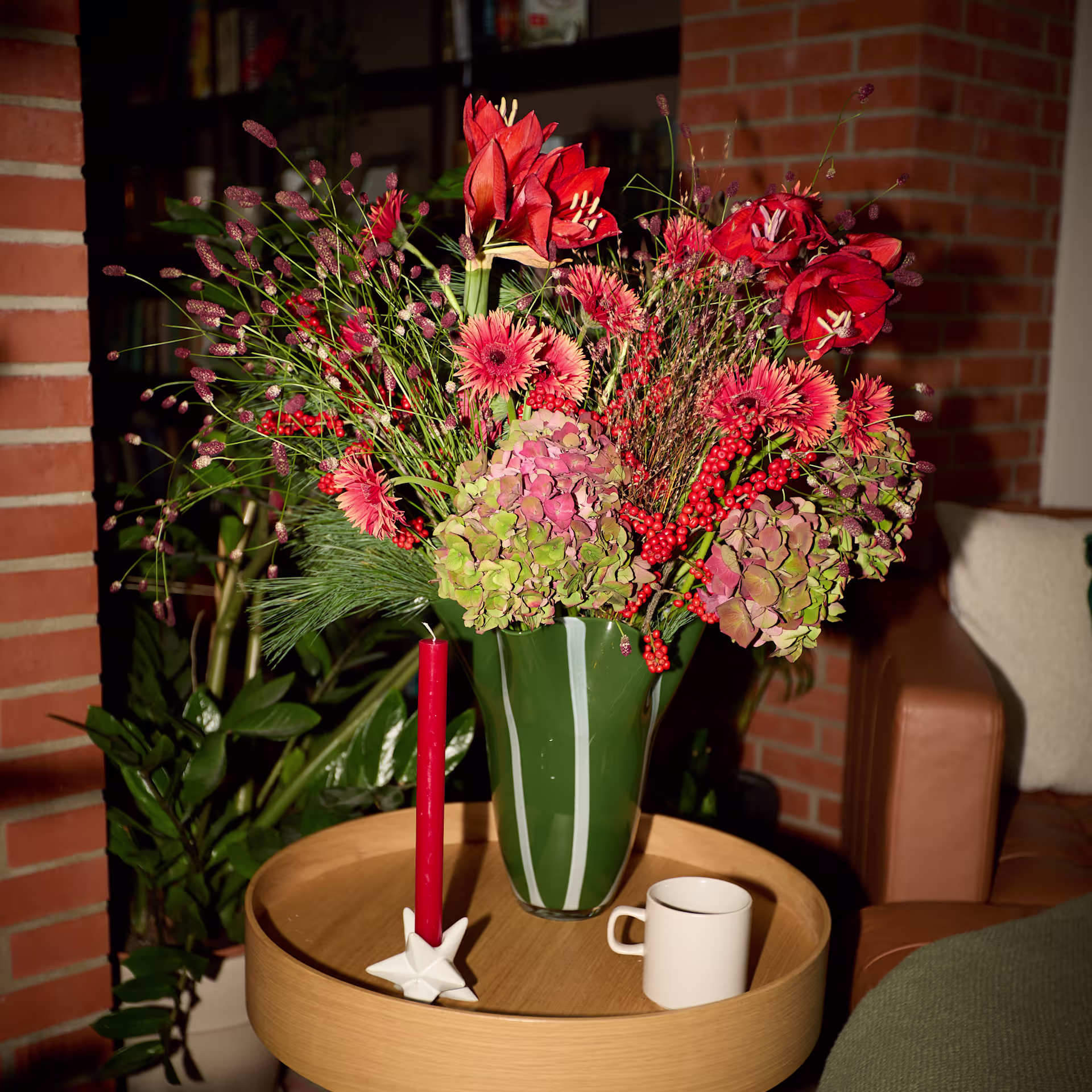 Auf einem Holztisch vor einer Backsteinmauer und einem Ledersofa stehen ein farbenfrohes Blumenarrangement in einer grünen Vase und eine rote Kerze. (Beschreibungstext KI-generiert)