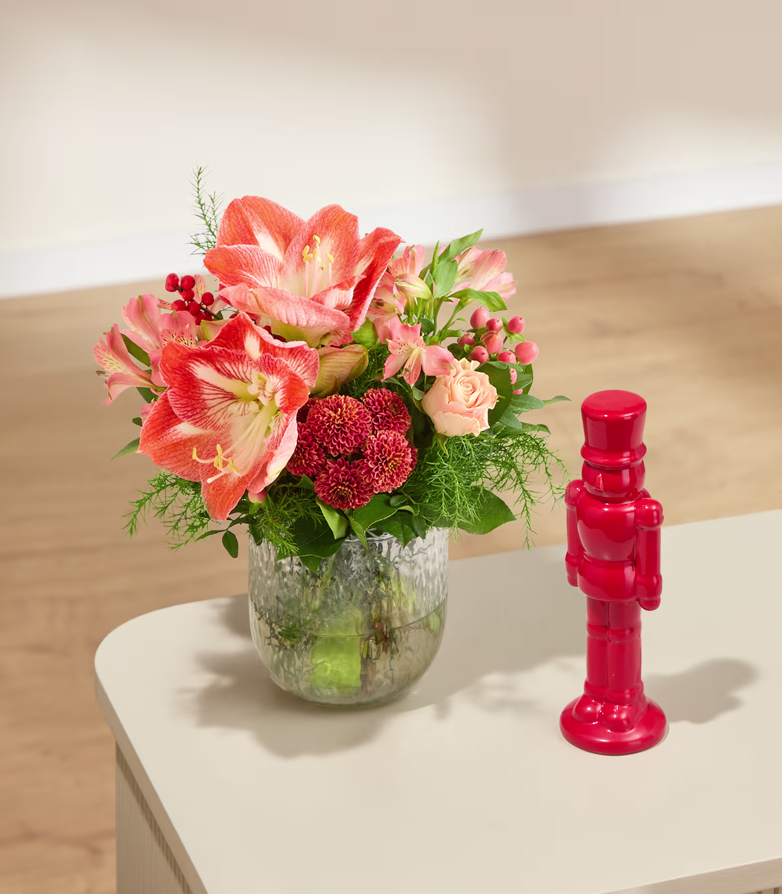 Auf einem weißen Tisch vor einer schlichten Wand und einem hellen Holzboden stehen ein Strauß orangefarbener und roter Blumen in einer Glasvase und eine rote Nussknackerfigur. (Beschreibungstext KI-generiert)