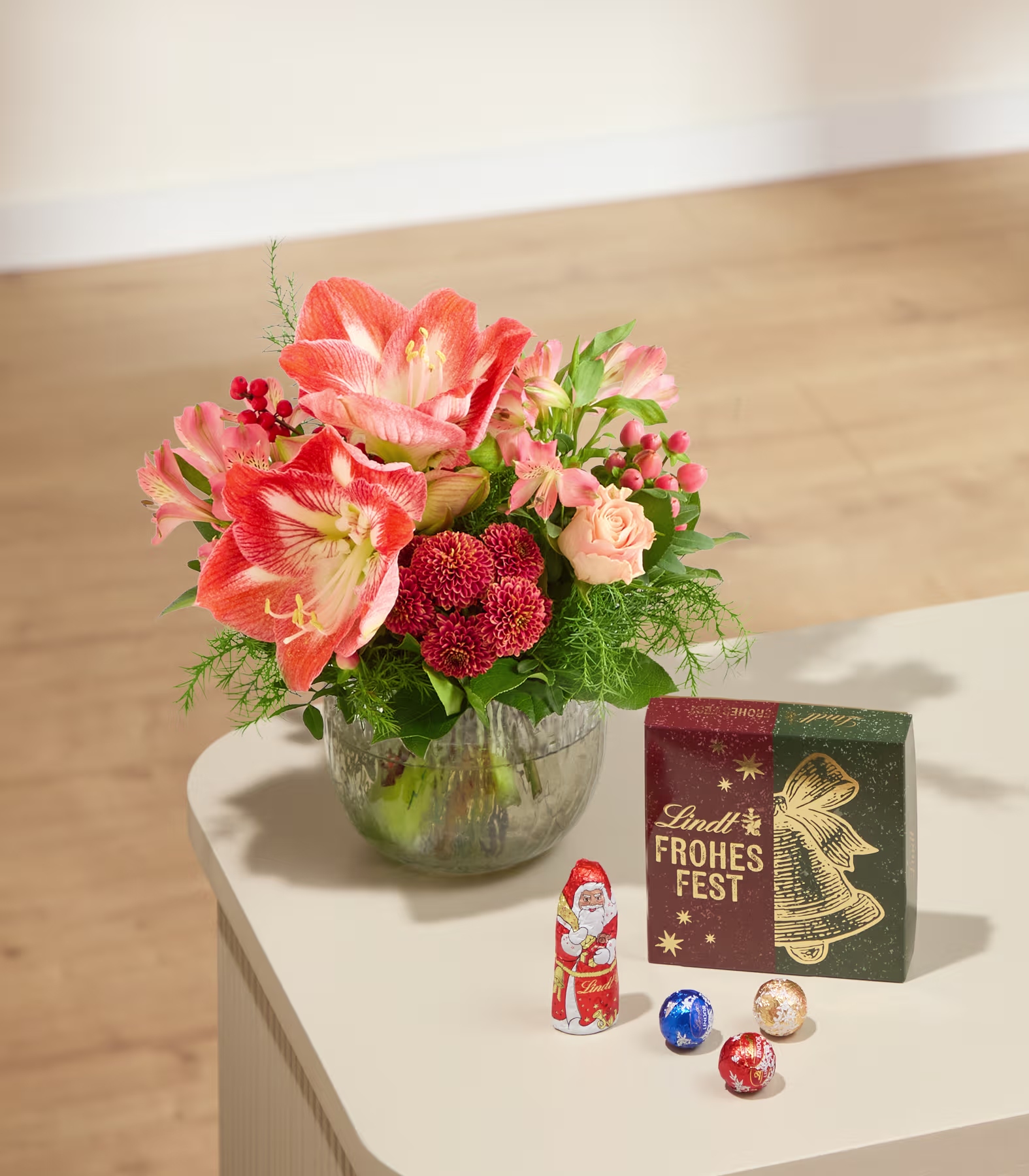 Auf einem weißen Tisch steht ein Strauß orangefarbener und roter Blumen in einer Glasvase neben einer Lindt-Pralinenschachtel und einer kleinen Weihnachtsmannfigur vor einem neutralen Innenraumhintergrund. (Beschreibungstext KI-generiert)