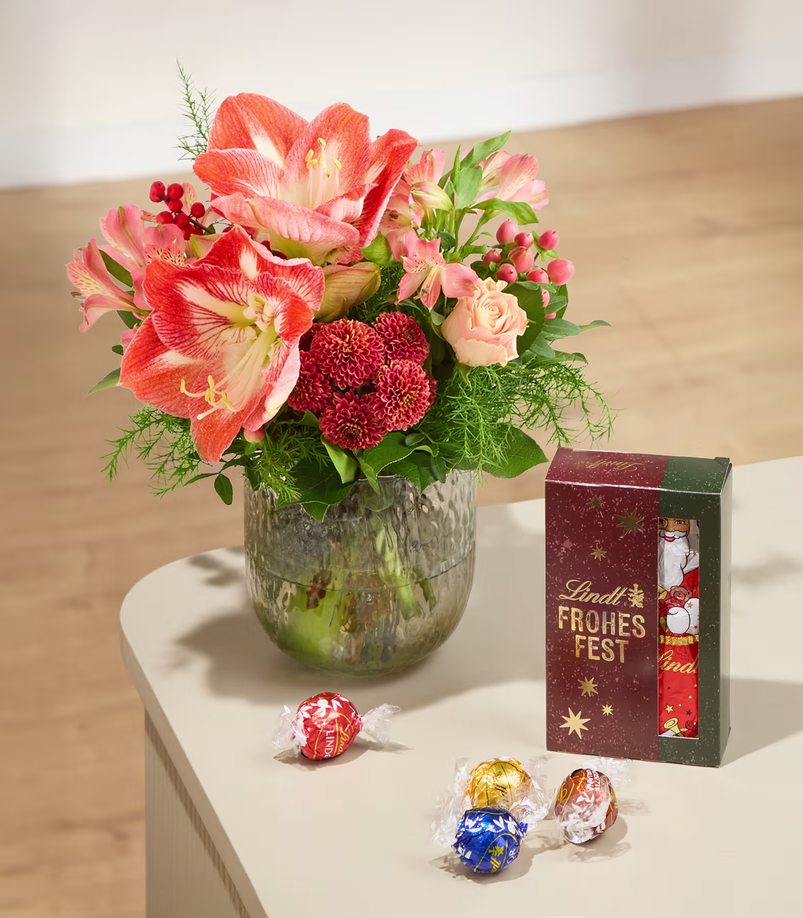 Ein leuchtender Strauß orangefarbener und roter Blumen in einer Glasvase steht neben einer Lindt-Schokoladengeschenkbox und verstreuten Trüffeln auf einem hellen Tisch vor neutralem Hintergrund. (Beschreibungstext KI-generiert)