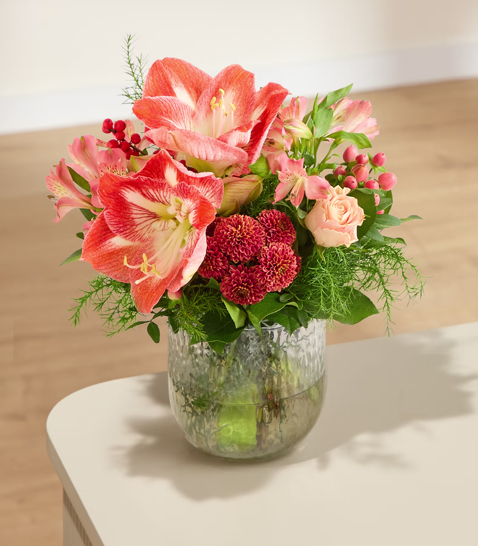 Ein leuchtender Strauß orangefarbener und rosafarbener Blumen in einer Glasvase steht auf einem weißen Tisch vor einem neutralen, minimalistischen Hintergrund. (Beschreibungstext KI-generiert)