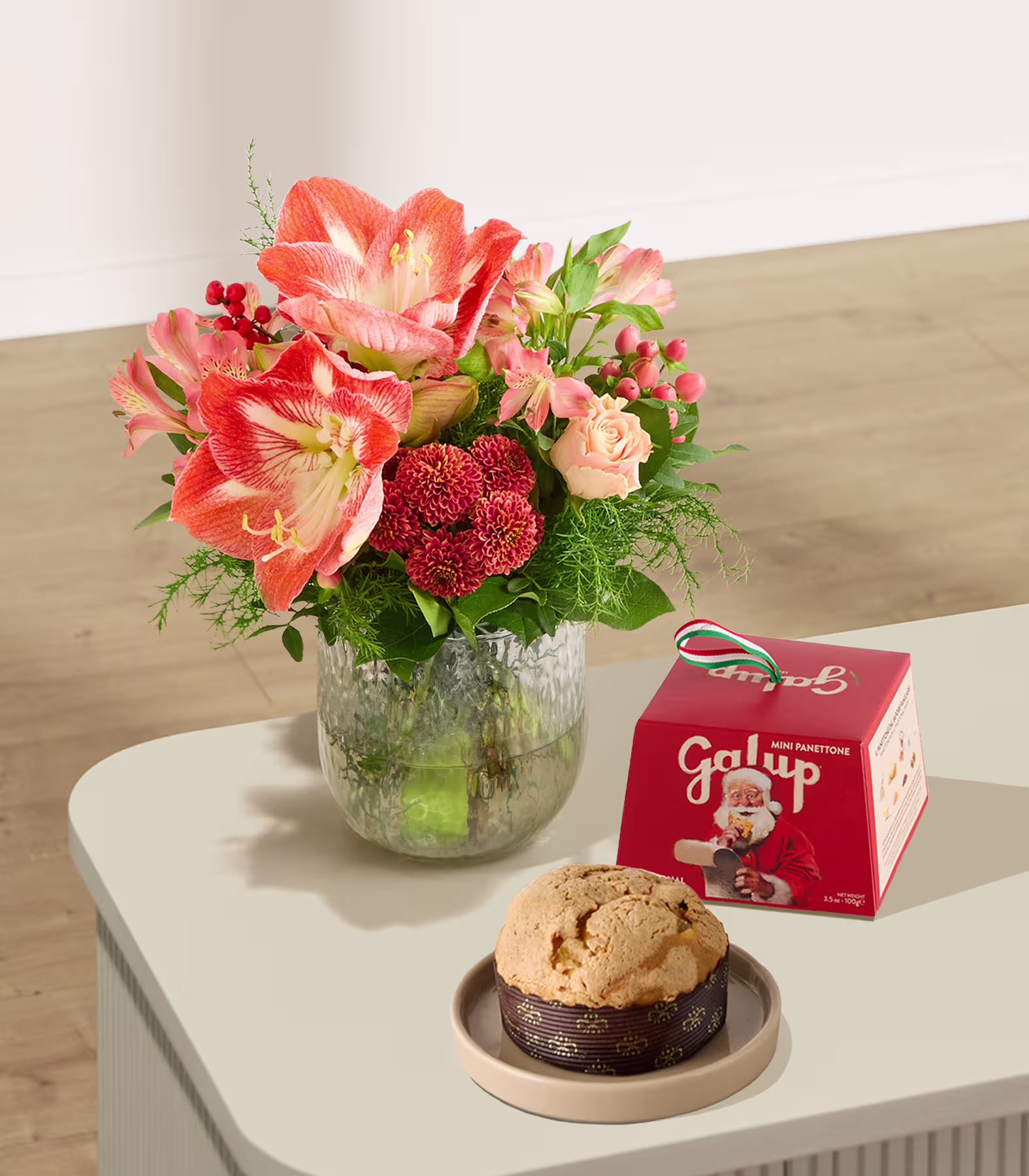 Auf einem Tisch steht ein farbenprächtiger Strauß roter und rosafarbener Blumen in einer Glasvase, daneben eine kleine Schachtel und ein einzelnes Gebäck auf einem Teller. (Beschreibungstext KI-generiert)