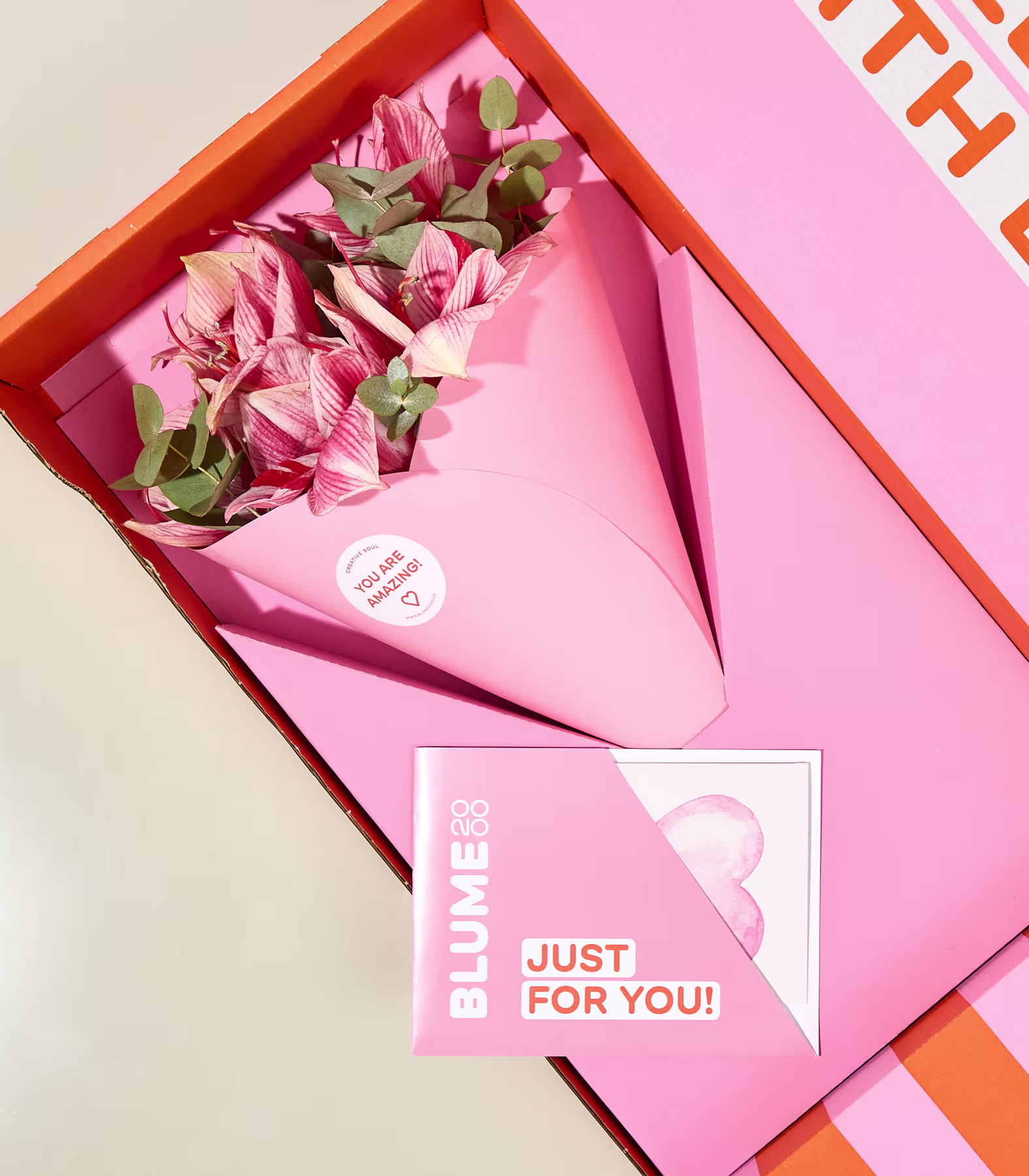 In einer leuchtend orange-rosa Geschenkbox befinden sich ein Strauß rosa Blumen, eingewickelt in rosa Papier, und eine Karte mit der Aufschrift „NUR FÜR DICH!“. (Beschreibungstext KI-generiert)