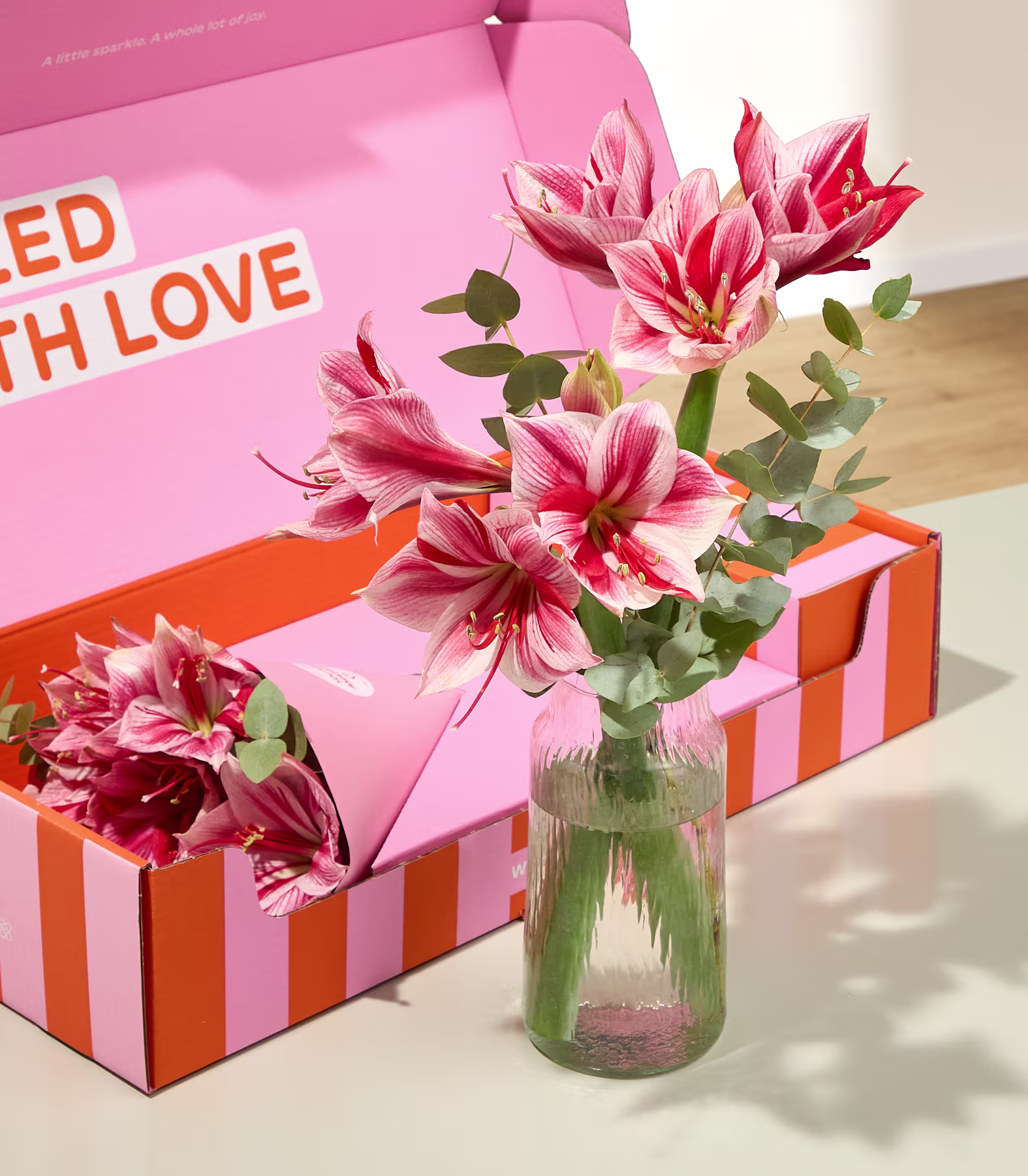 Im Vordergrund stehen rosa und rote Lilien in einer Glasvase, im Hintergrund eine dazu passende rosa-orangefarbene Geschenkbox. (Beschreibungstext KI-generiert)