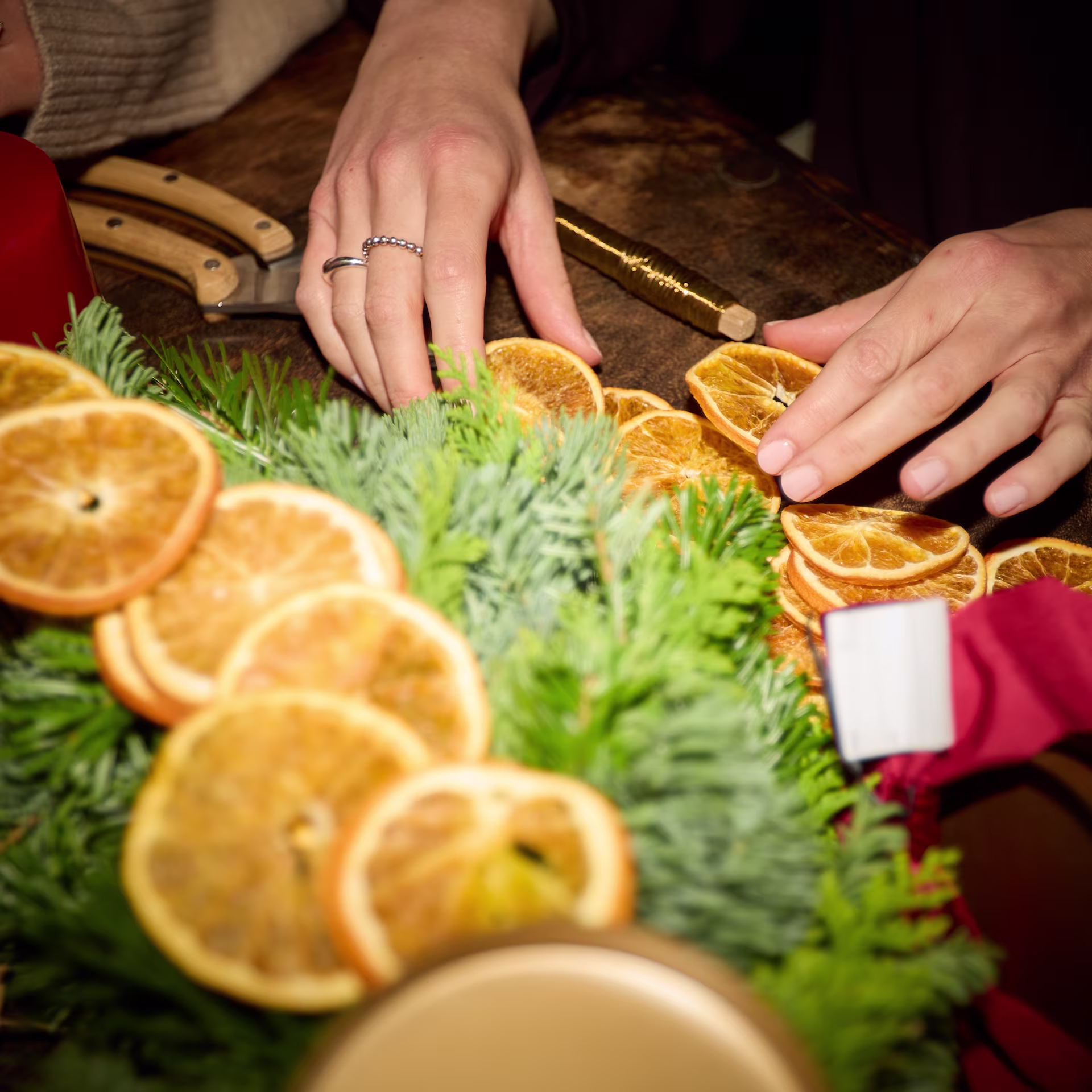 Im Vordergrund liegen getrocknete Orangenscheiben in einem grünen immergrünen Kranz, während im Hintergrund die Hände einer Person auf einem Holztisch mit weiteren Orangenscheiben und Bastelwerkzeugen arbeiten. (Beschreibungstext KI-generiert)