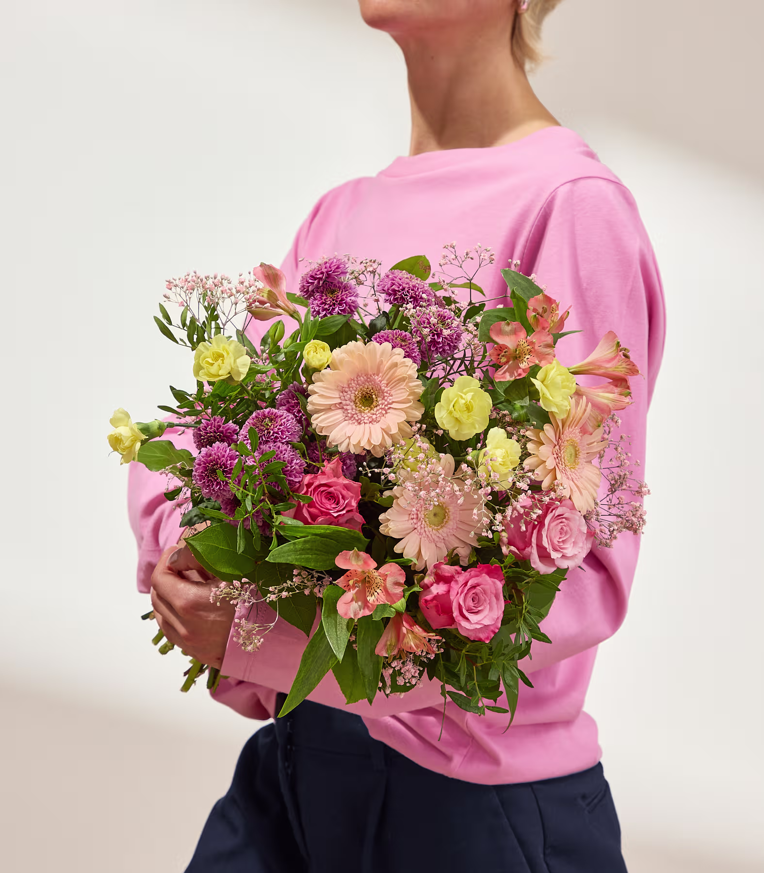 Eine lächelnde Frau in einem hellrosa Hemd hält einen farbenfrohen Strauß aus rosa, gelben und pfirsichfarbenen Blumen mit zartem weißen Schleierkraut vor einem sanft beleuchteten, cremeweißen Hintergrund. (Beschreibungstext KI-generiert)