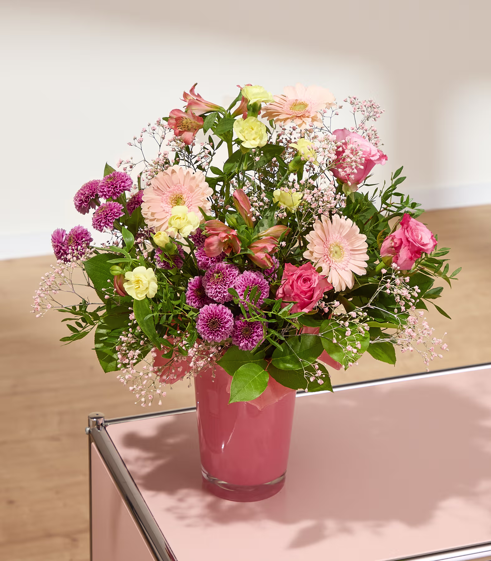 Im Vordergrund steht ein farbenprächtiger Blumenstrauß in einer Glasvase auf einem runden Holztisch, im Hintergrund ein sonnenbeschienenes Fenster und ein gemütliches Sofa. (Beschreibungstext KI-generiert)