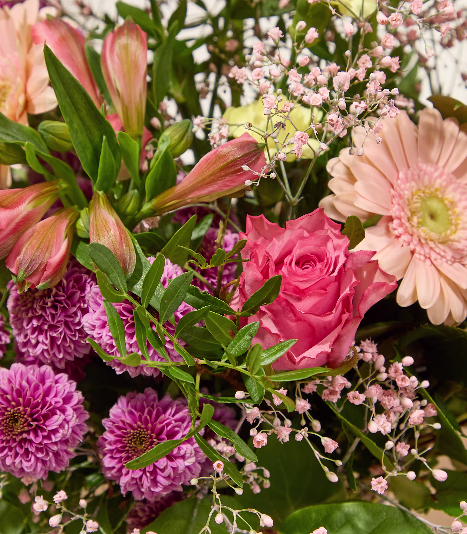 Eine Person in einem hellrosa Hemd hält einen farbenfrohen Strauß aus rosa, gelben und violetten Blumen vor einem sanft beleuchteten, neutralen Hintergrund. (Beschreibungstext KI-generiert)