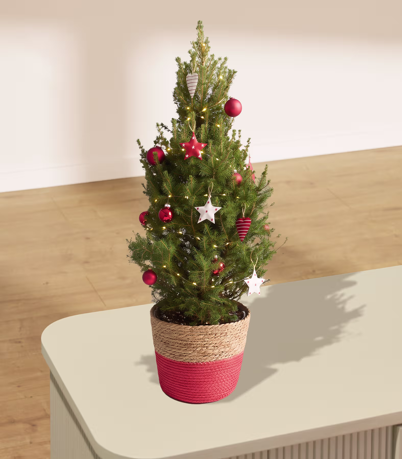 Ein kleiner, geschmückter Weihnachtsbaum in einem rot-braunen Flechttopf steht auf einem hellen Tisch vor einer schlichten weißen Wand und einem Holzboden. (Beschreibungstext KI-generiert)