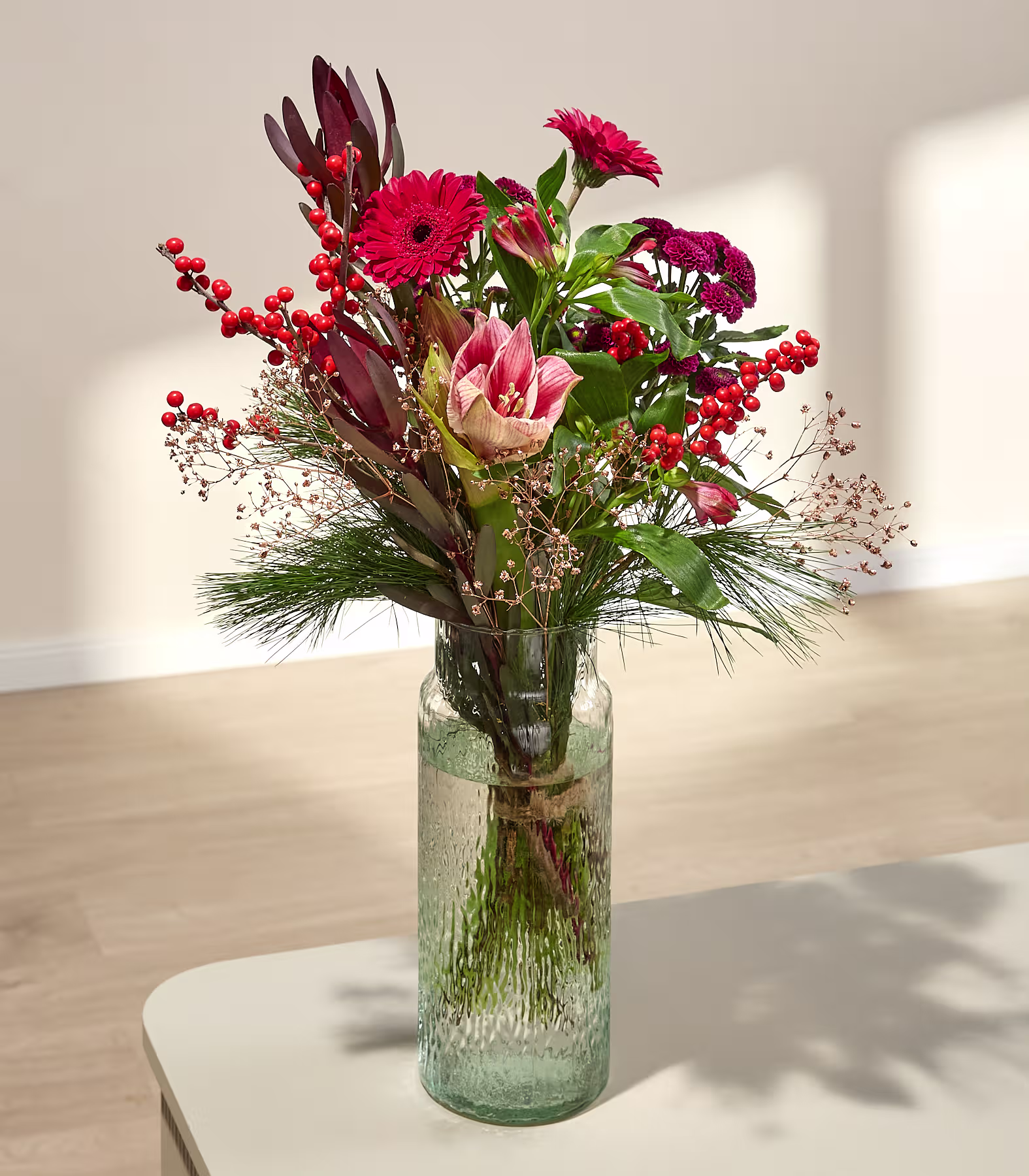 Ein leuchtend roter Blumenstrauß mit Beeren in einer durchsichtigen Glasvase steht auf einem hellen Tisch vor einer neutralen Wand und einem Holzboden. (Beschreibungstext KI-generiert)