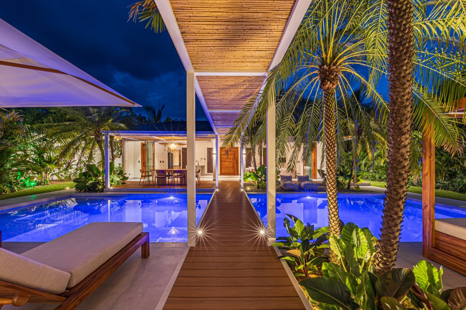 Wooden walkway between two illuminated pools in a tropical villa at night.