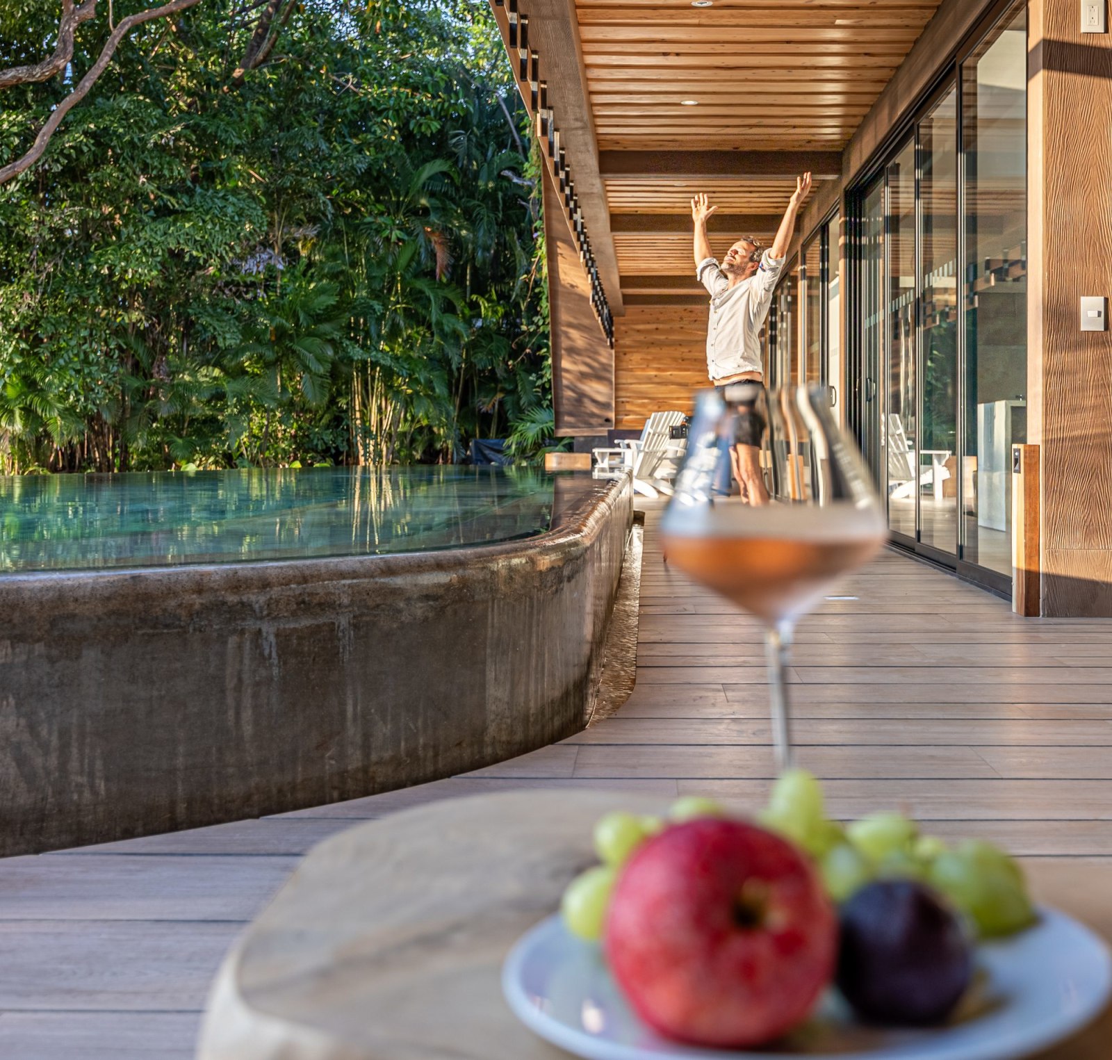 Man raises his arms joyfully on a terrace with a wine glass and fruit in foreground.