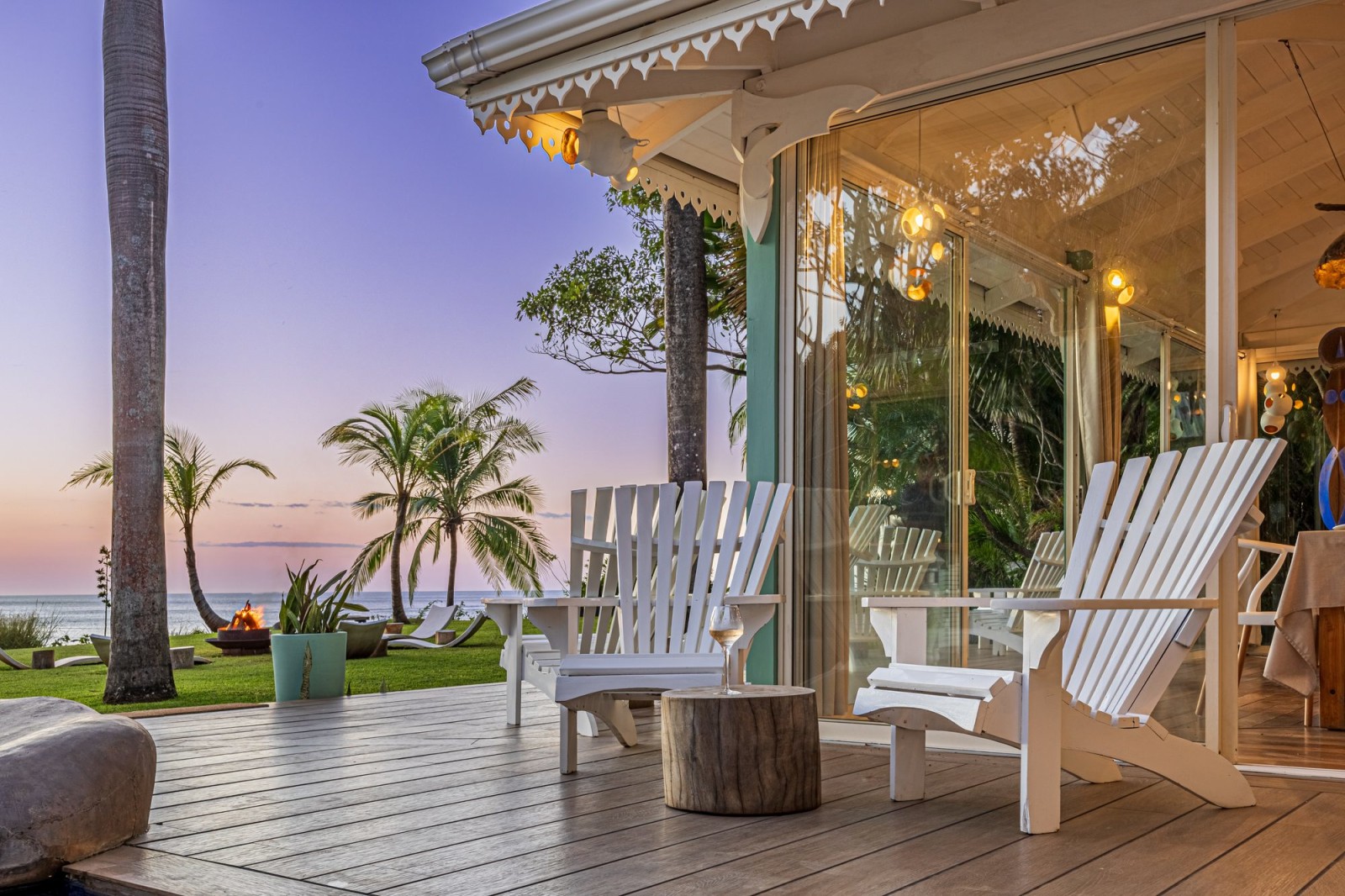 Wooden deck with white chairs facing the ocean at sunset among palm trees.
