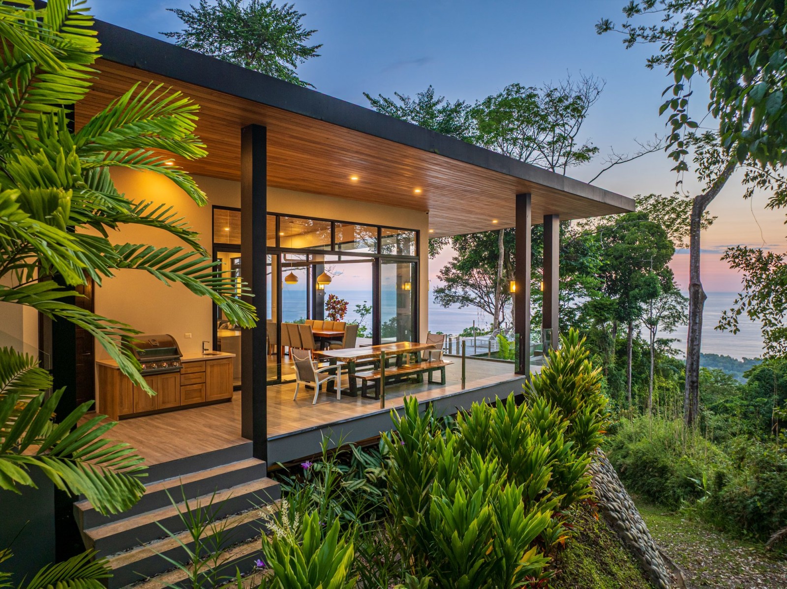 Covered terrace with outdoor dining and ocean view through tropical greenery at sunset