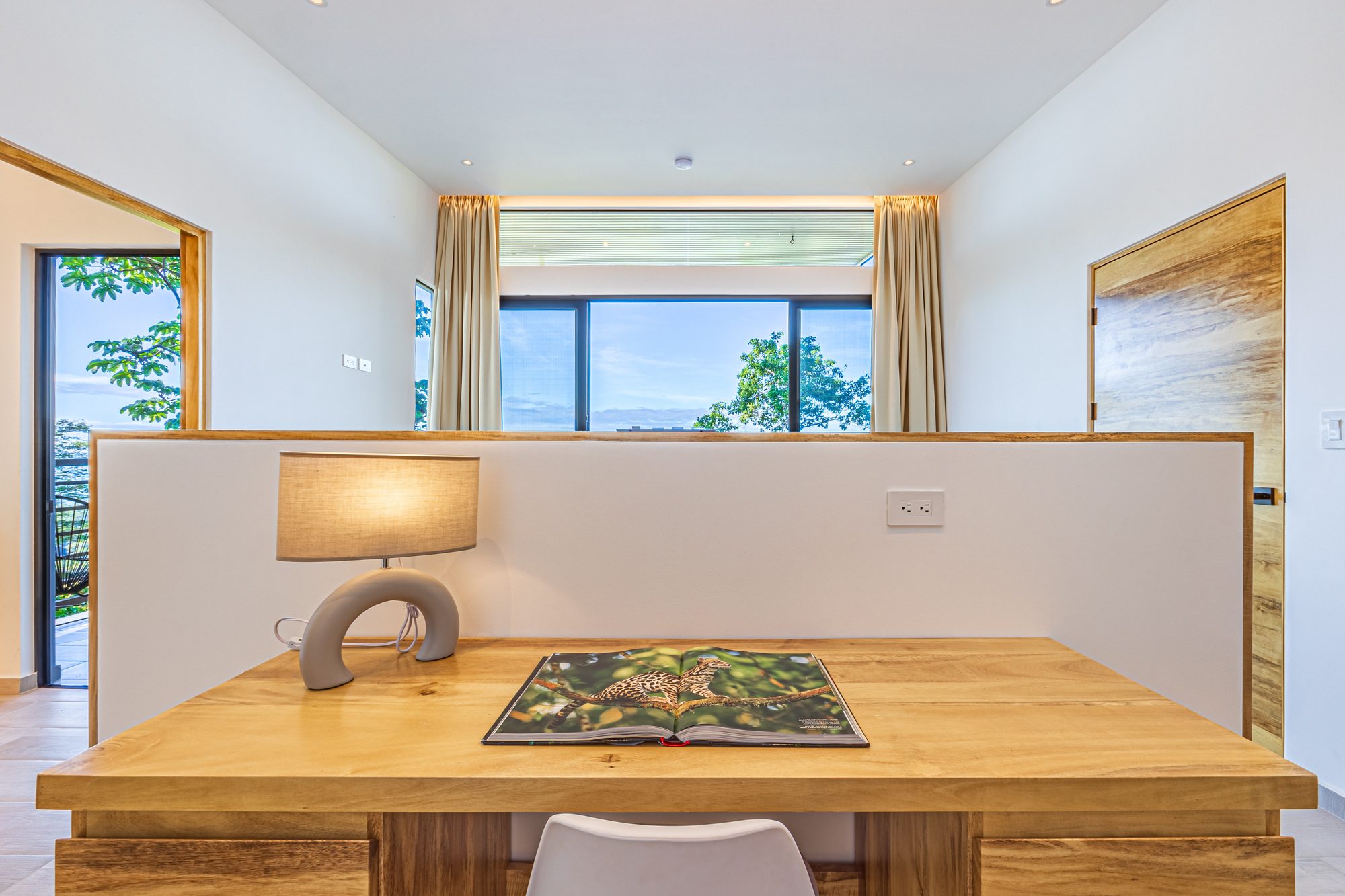 Wooden desk with lamp and open book facing a window with tree views.