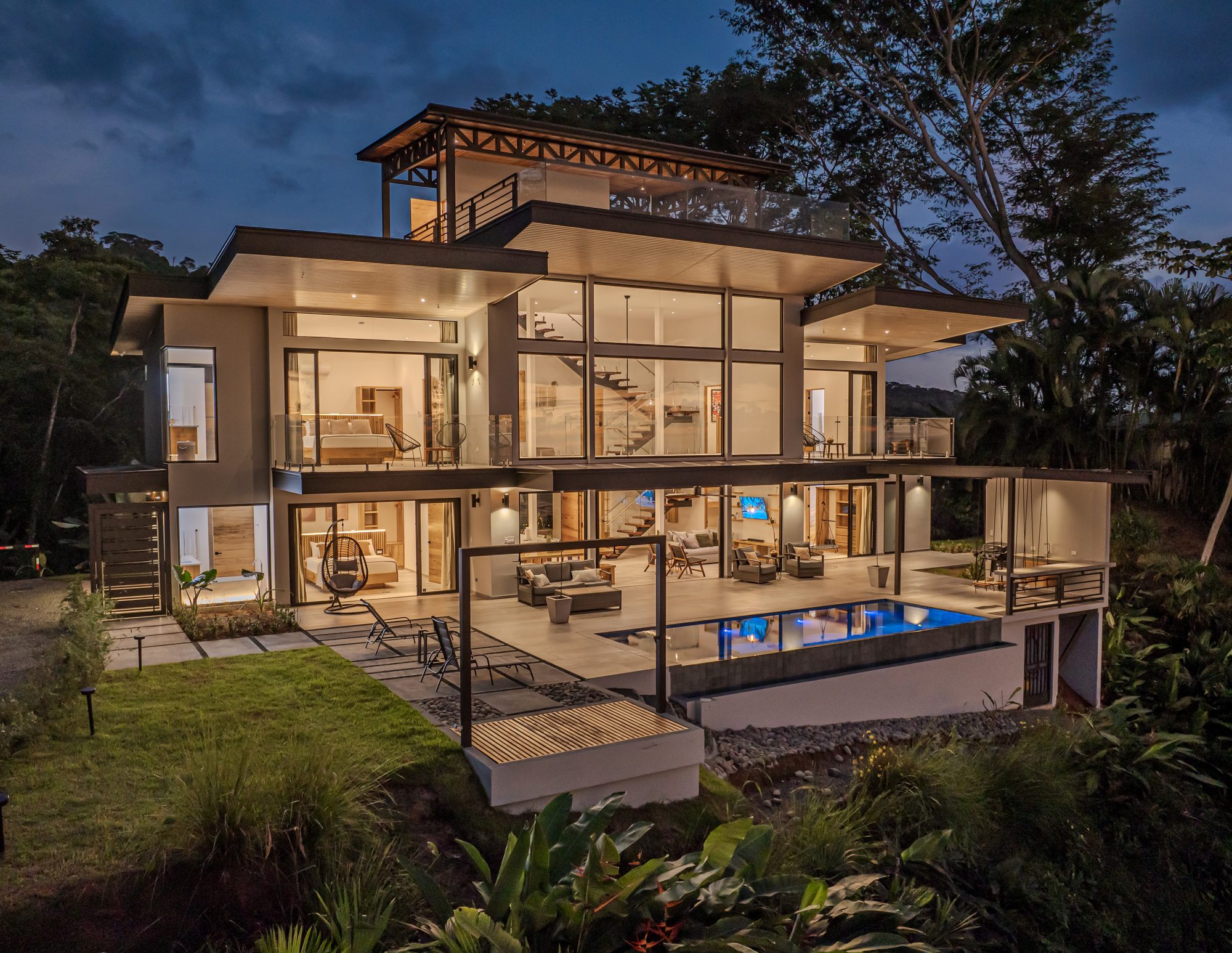 Night view of a modern three-story house with pool and tropical garden.