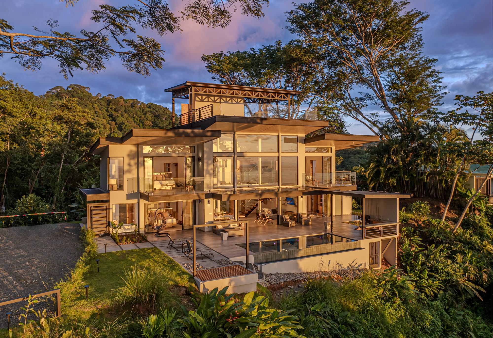 General view of a modern three-story villa surrounded by jungle at sunset.