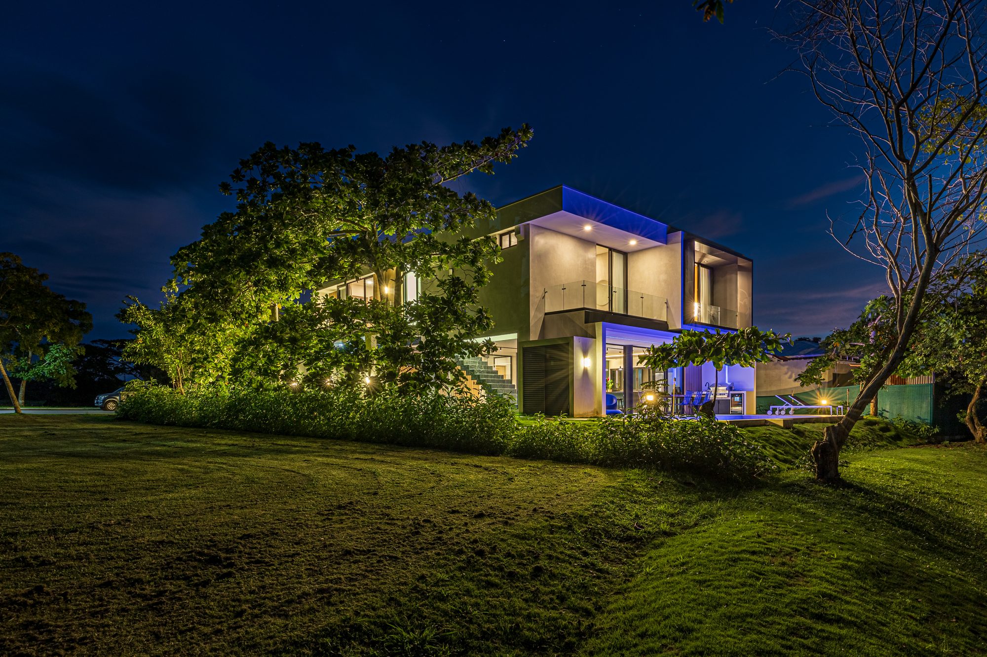 Night view of a modern two-story house with blue lighting and a large garden.