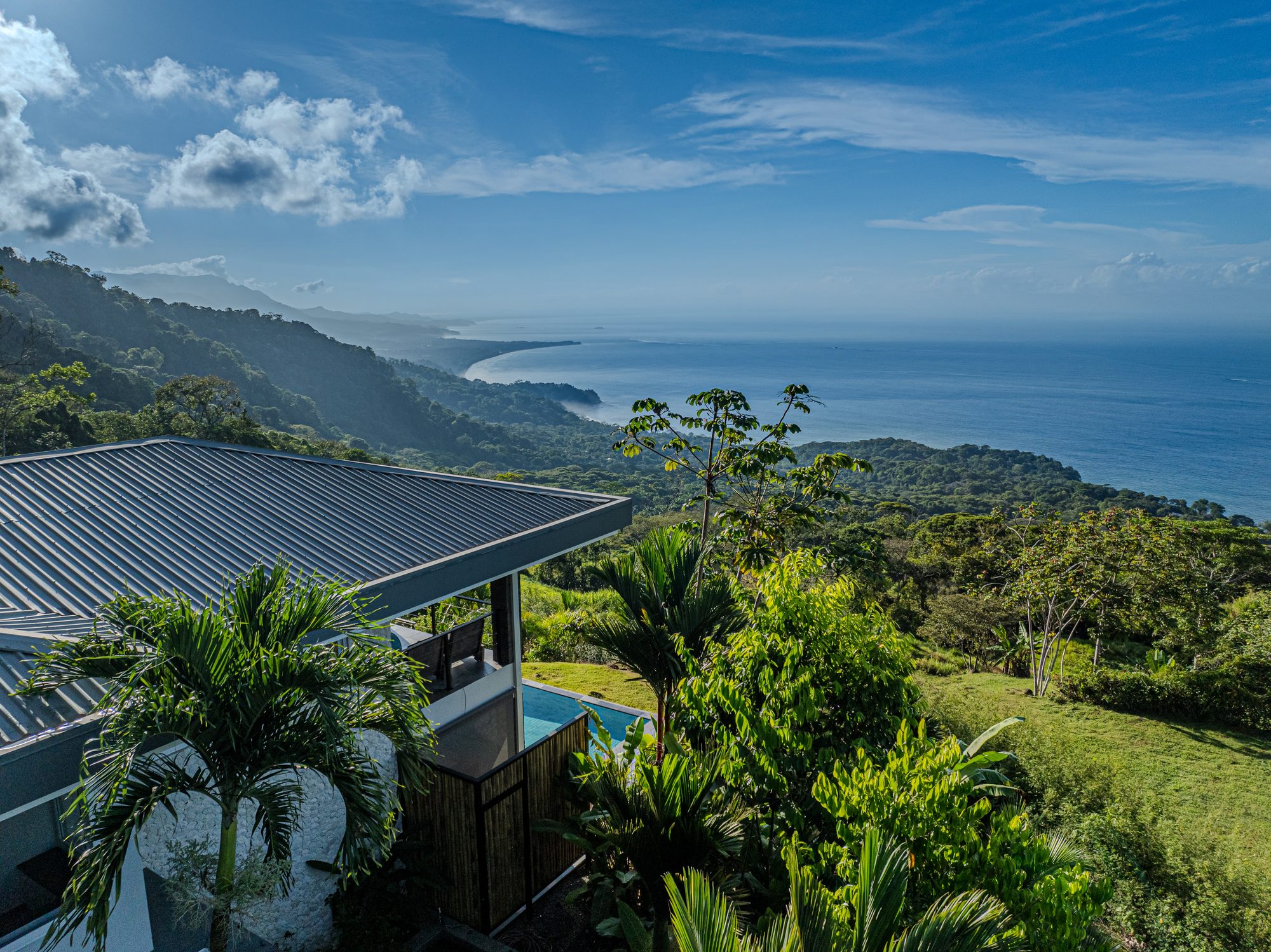 Aerial view of a house with an infinity pool surrounded by jungle and the ocean beyond.