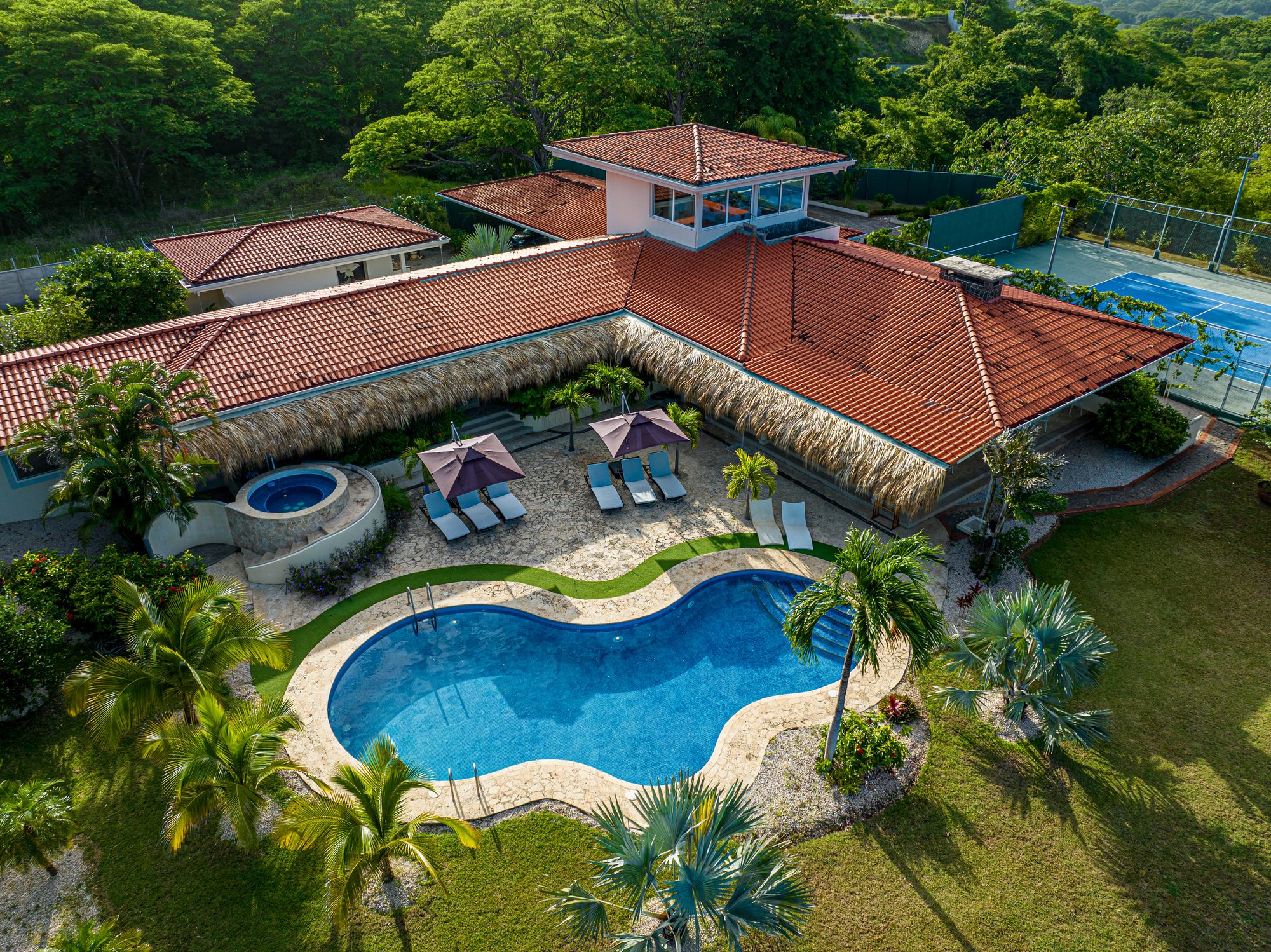Aerial view of a luxury villa with pool, jacuzzi and tennis court surrounded by jungle.