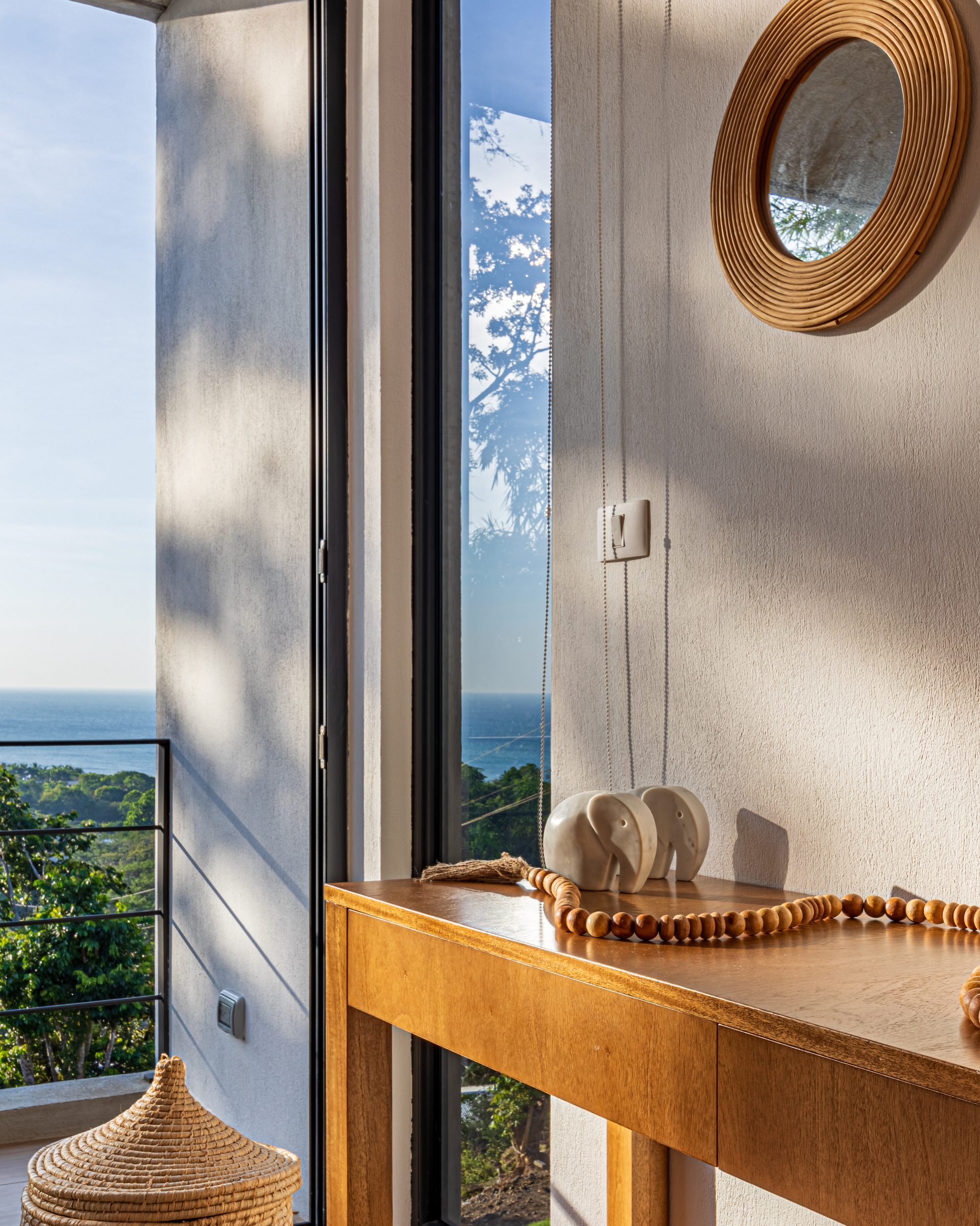 Wooden console table with decor near a window overlooking the ocean and a round rattan mirror.