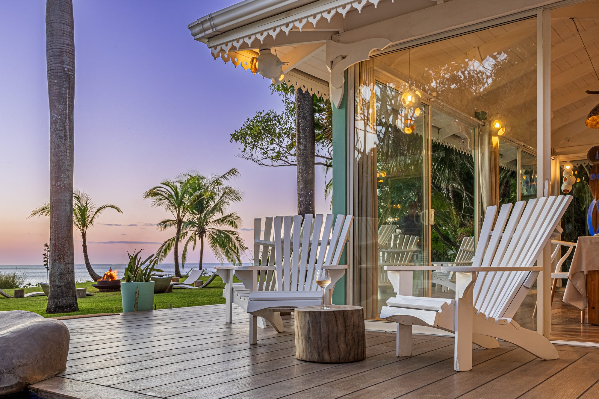 Terraza de madera con sillas blancas frente al mar al atardecer entre palmeras.