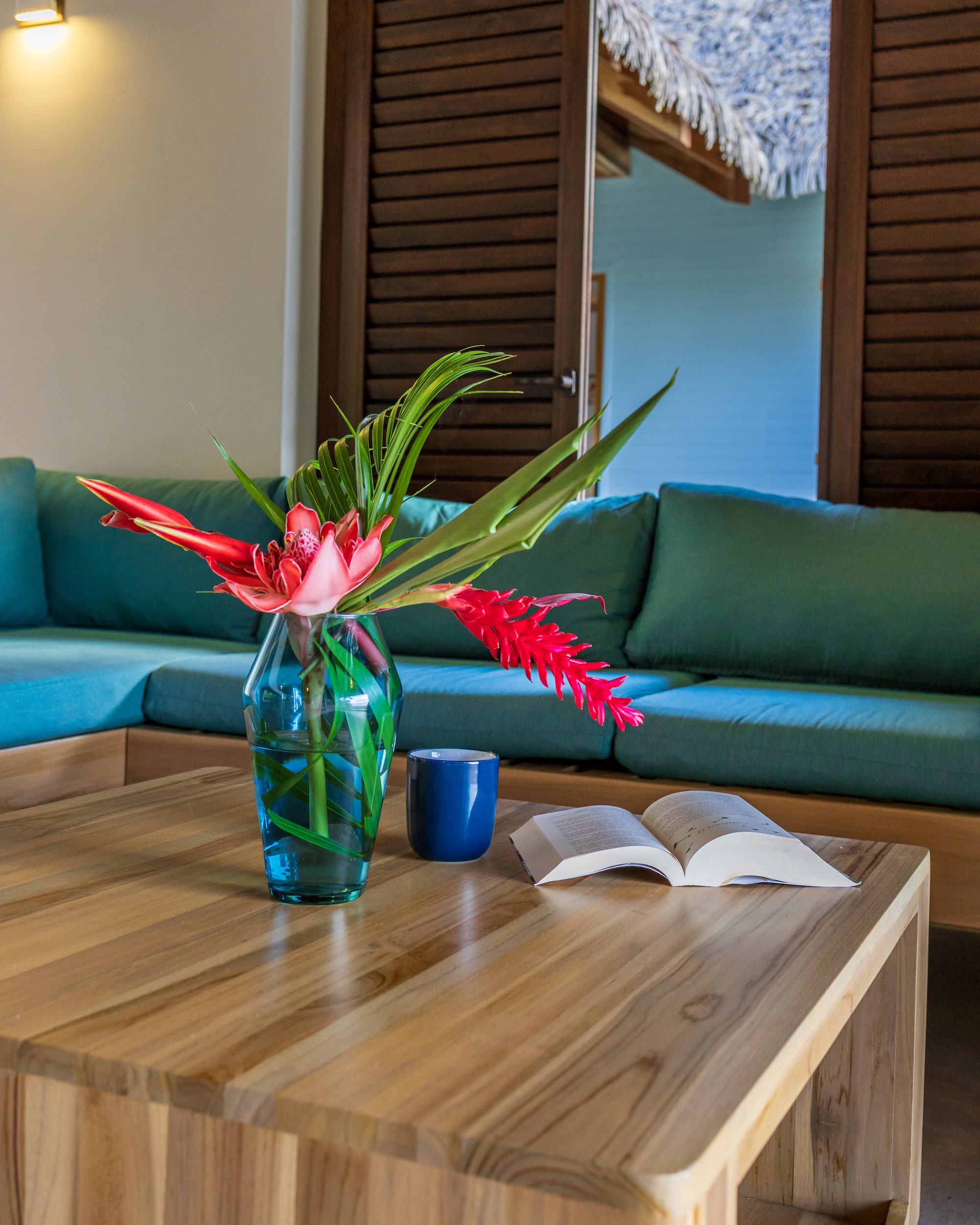 Wooden table with tropical flowers, blue mug and open book in front of green sofa.