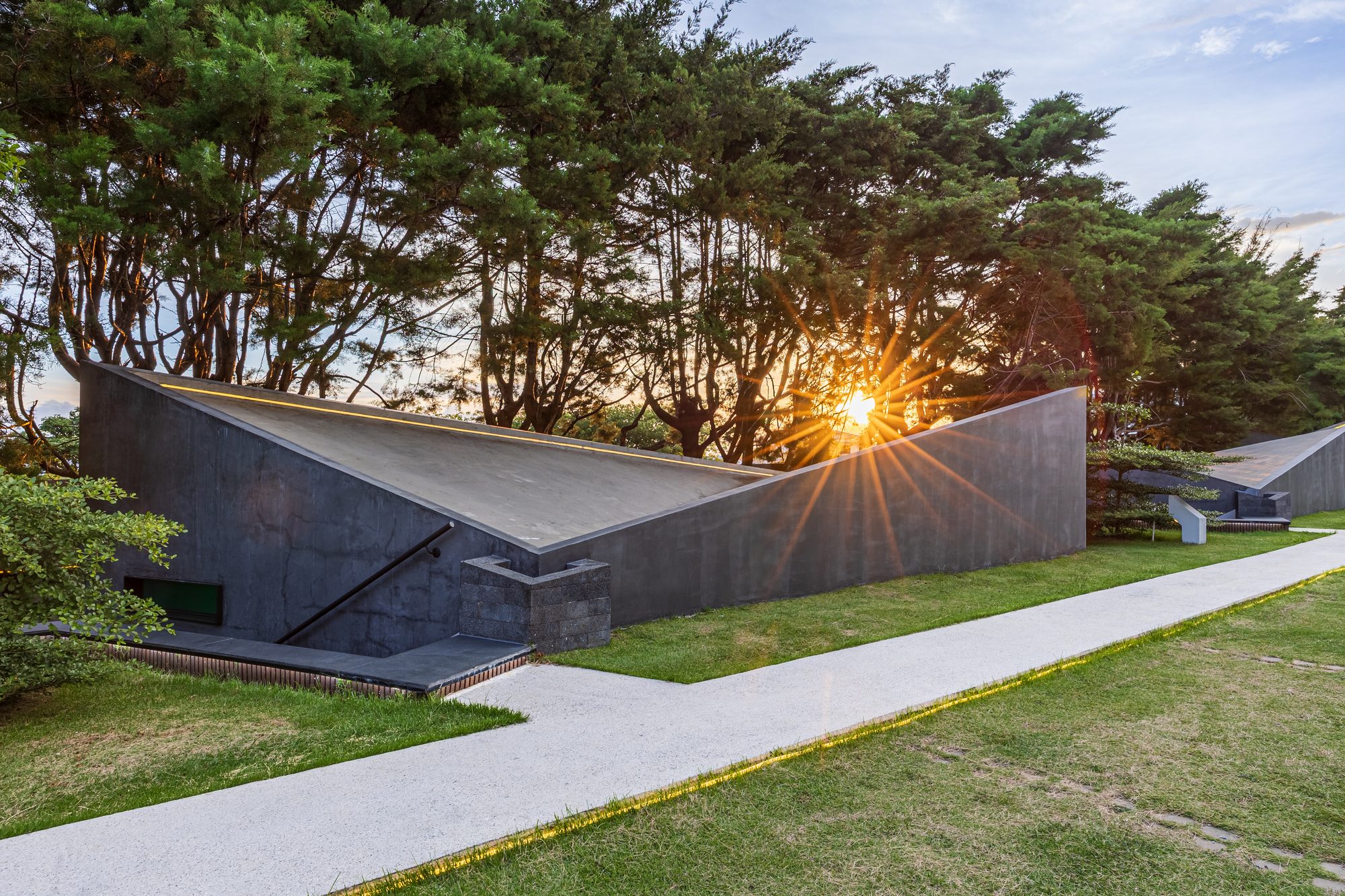 Angular concrete roof with sunburst through trees at sunset.