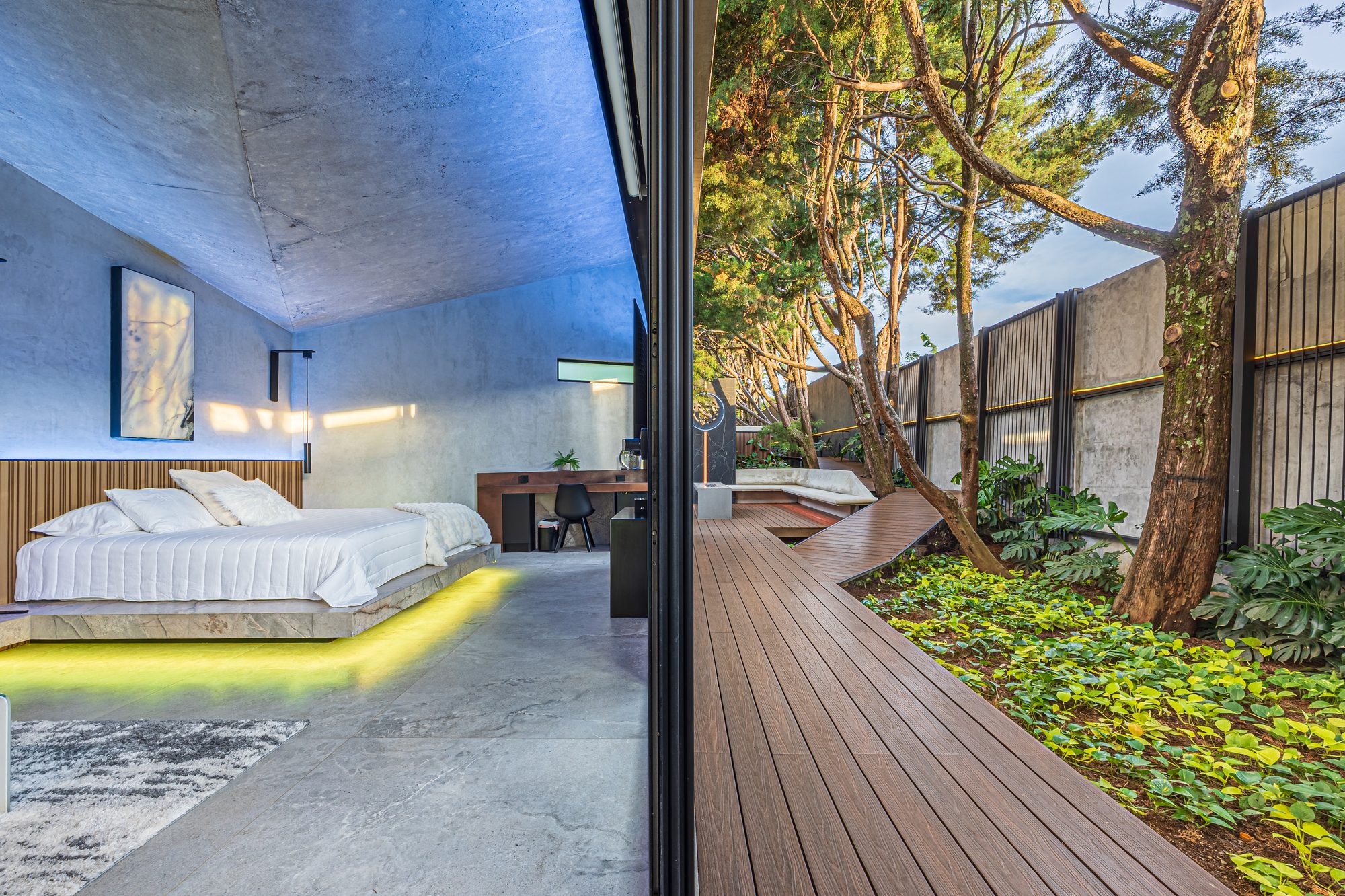Modern bedroom with curved concrete ceiling opening to a wooden deck among trees.