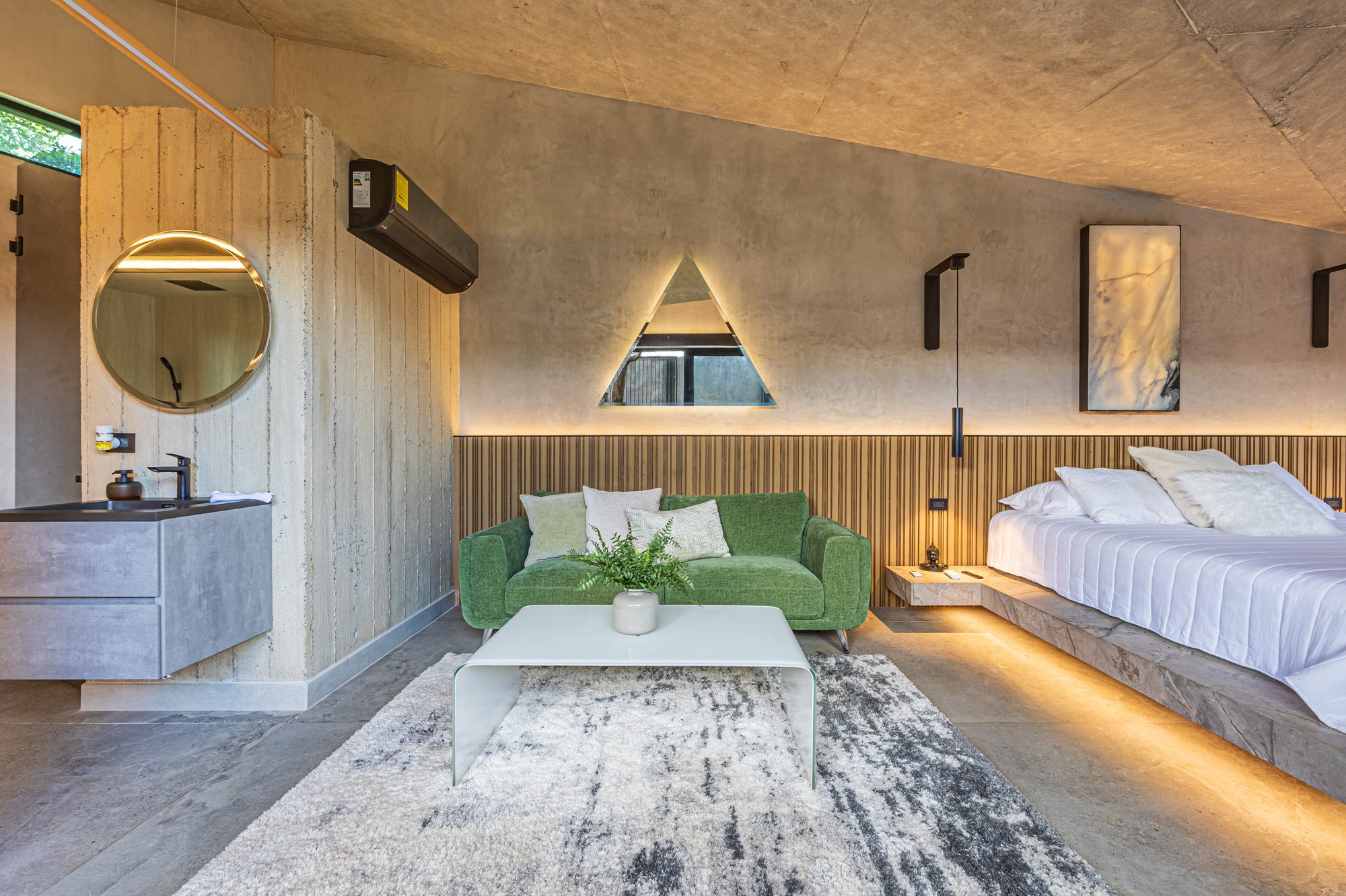 Suite with green sofa, bed, and illuminated triangular window against textured concrete walls.