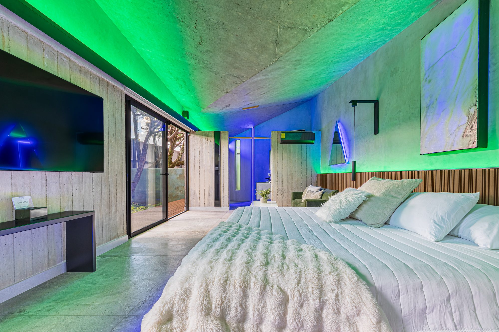 Bedroom with green and blue LED lighting, white bed and exposed concrete.