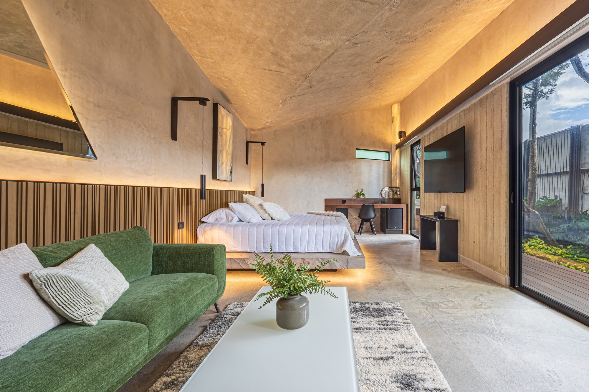 Suite with green sofa, floating bed and garden view through glass doors.