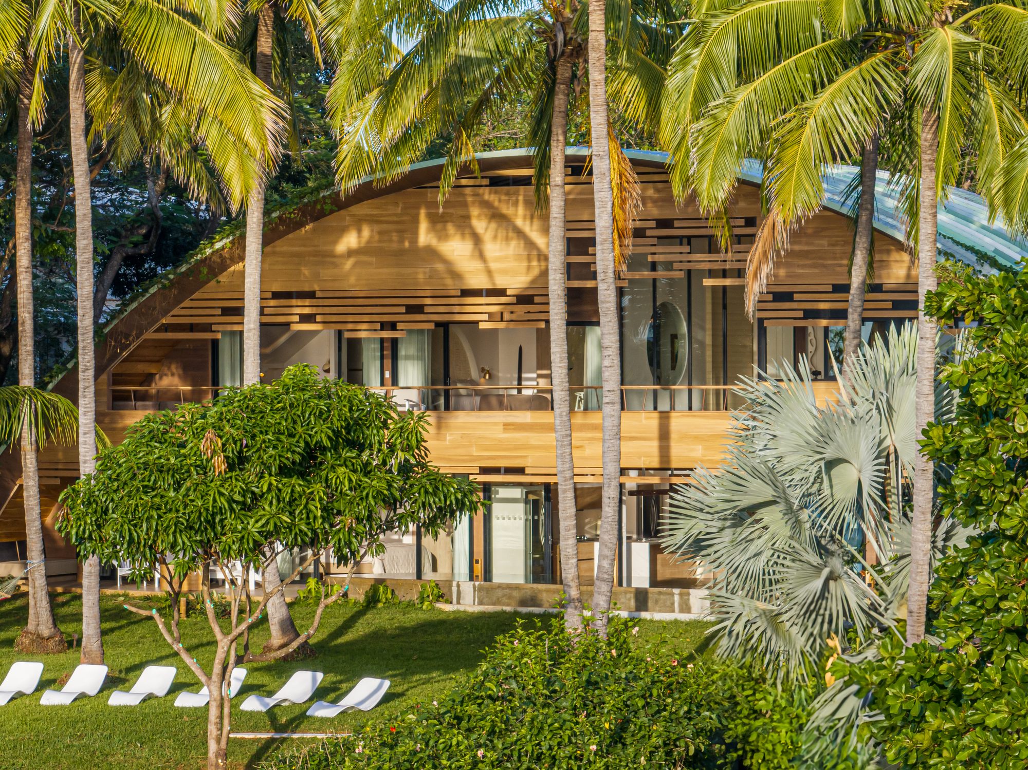 Two-story house with curved wood roof surrounded by tropical palm trees.