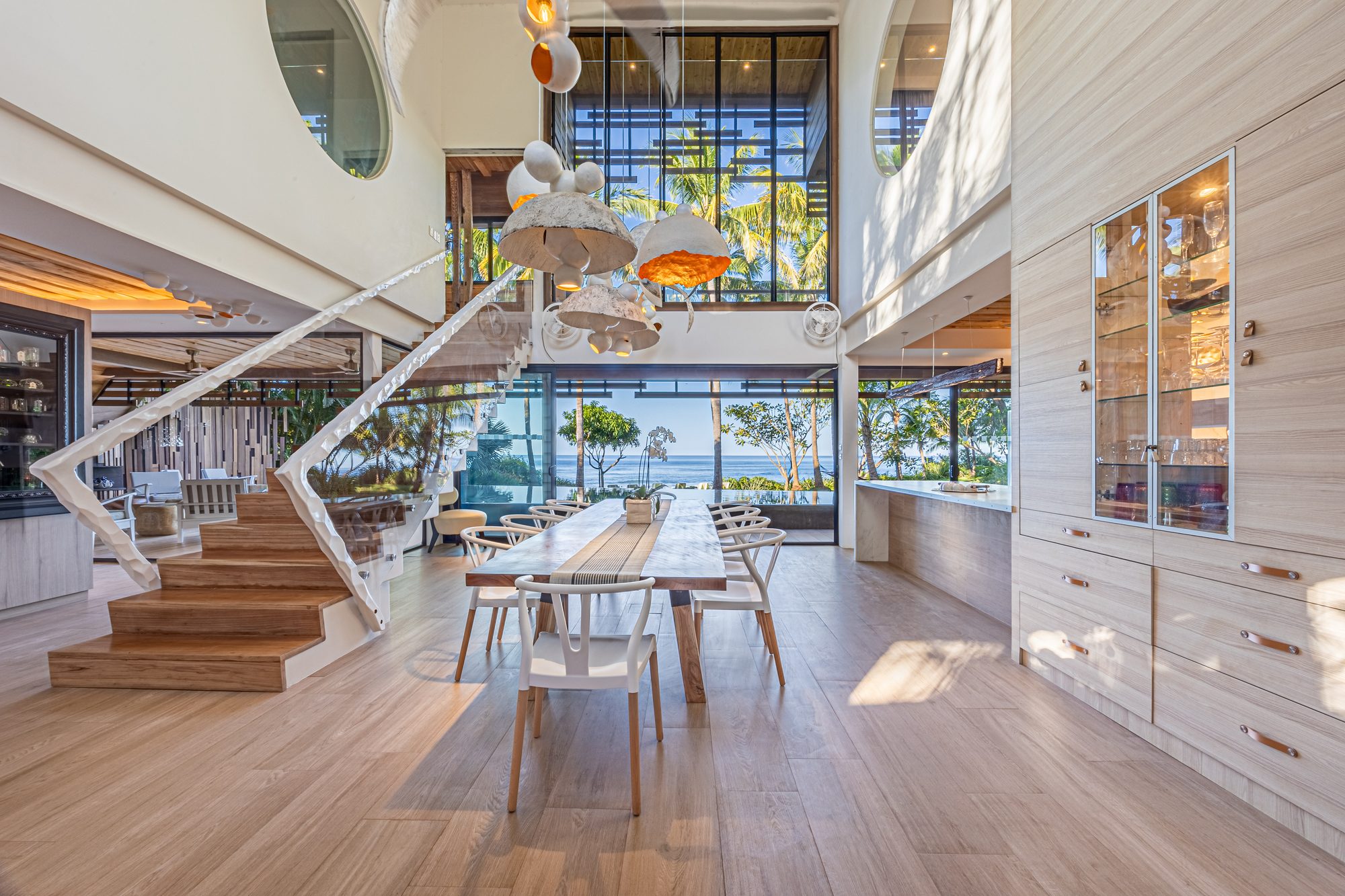 Double-height dining area with sculptural white staircase and ocean view through palms.