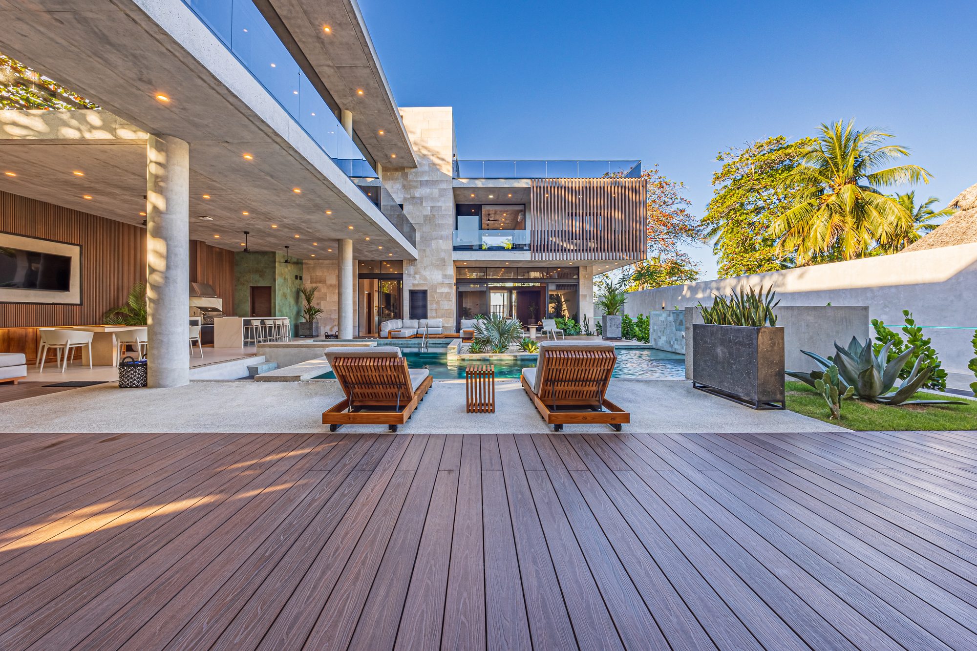 Wooden deck with pool and loungers in front of a modern house with palm trees.