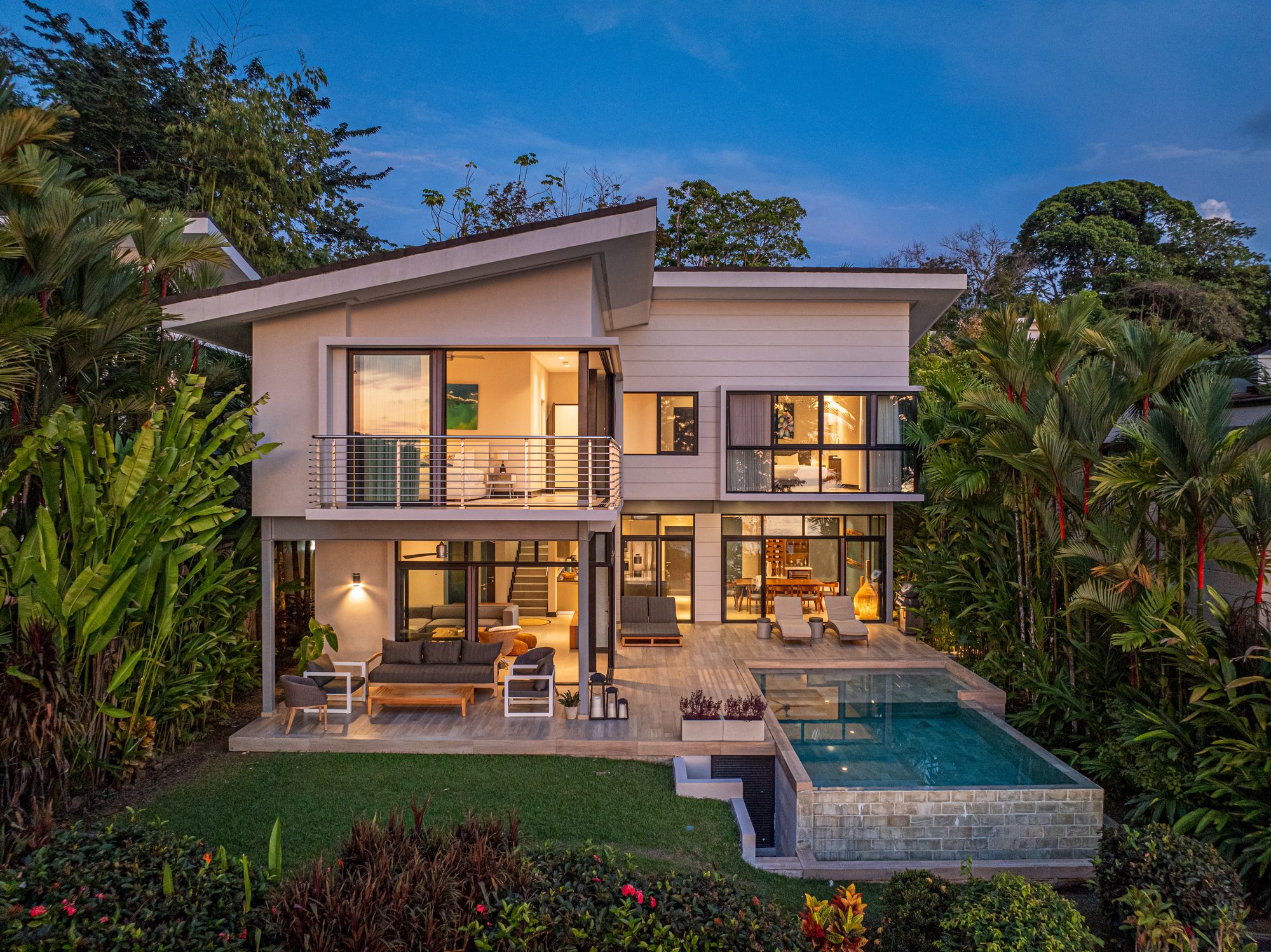 Modern two-story house with infinity pool at dusk surrounded by tropical greenery.