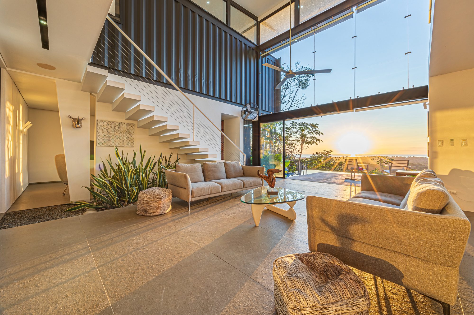 Living room with floating staircase and glass walls framing an ocean sunset.