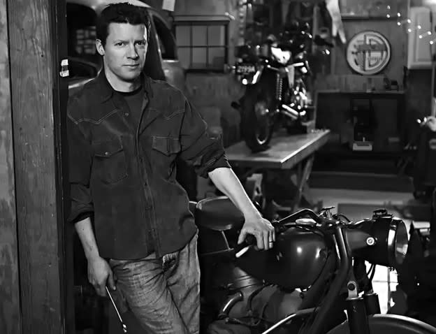 Black and white photograph of Matthew Crawford standing beside a motorcycle in his repair shop