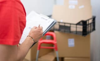 Un homme portant un tee-shirt rouge tient un presse-papiers et se tient devant des cartons empilés dans ce qui semble être une zone de stockage.